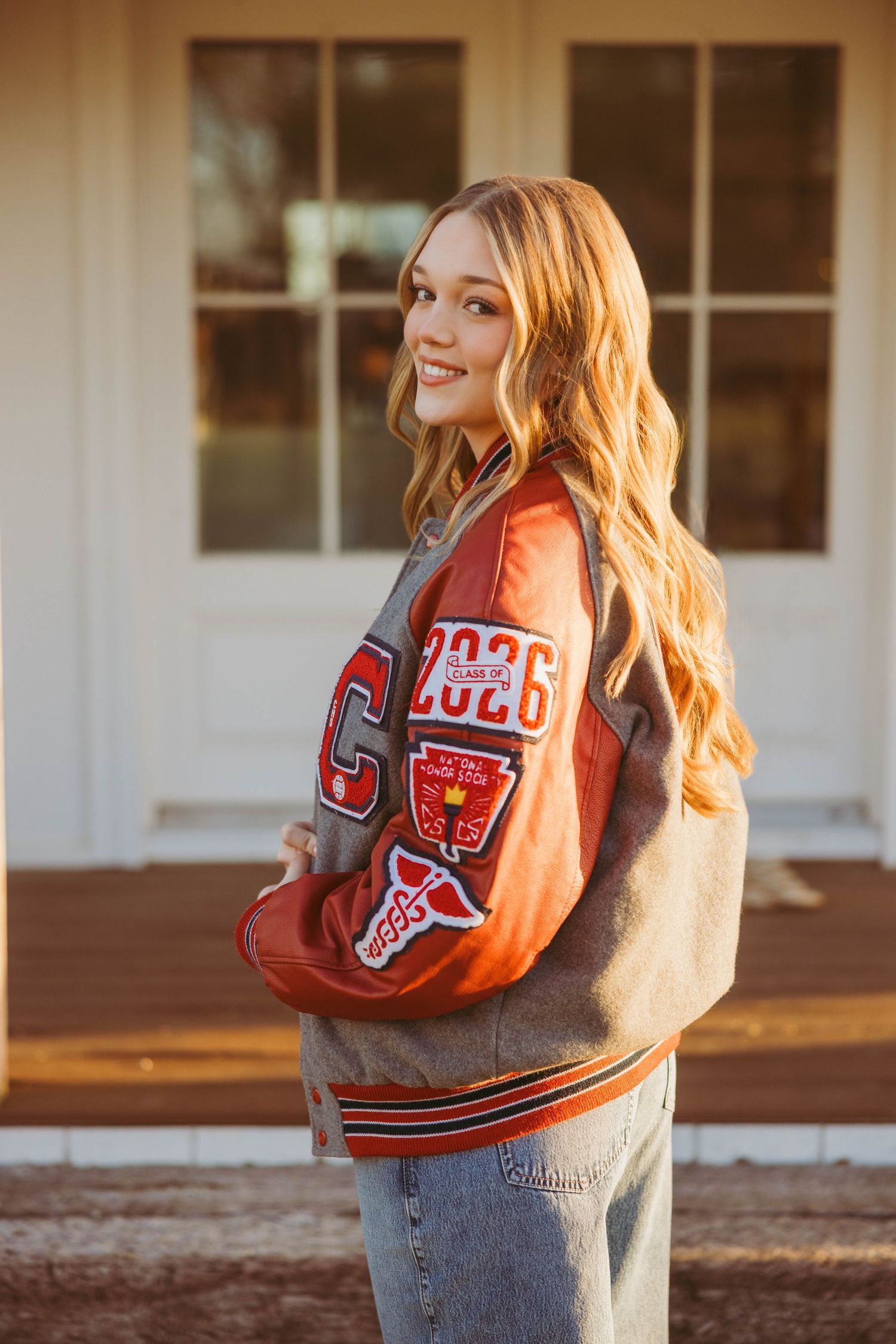 Smiling teenage girl wearing a letterman jacket with 2026 patches poses outdoors in warm sunlight.