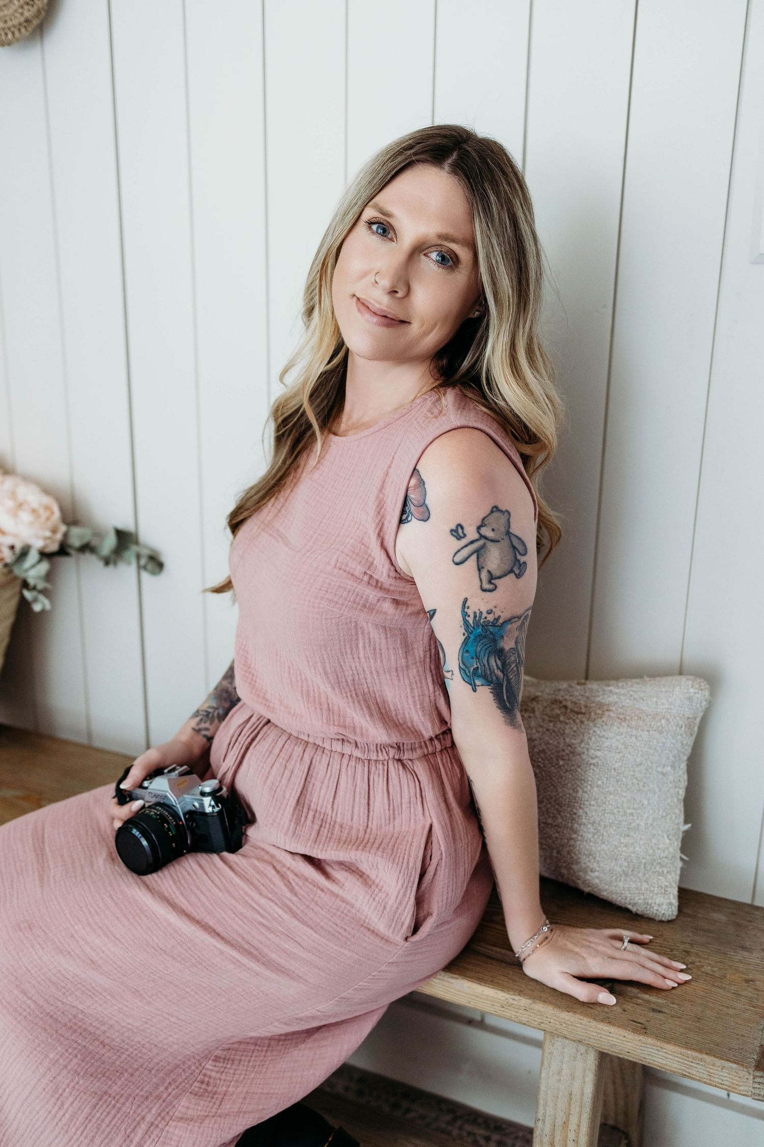 Woman in pink dress sitting on bench holding camera with tattoos on her arm, posing for a portrait photo.