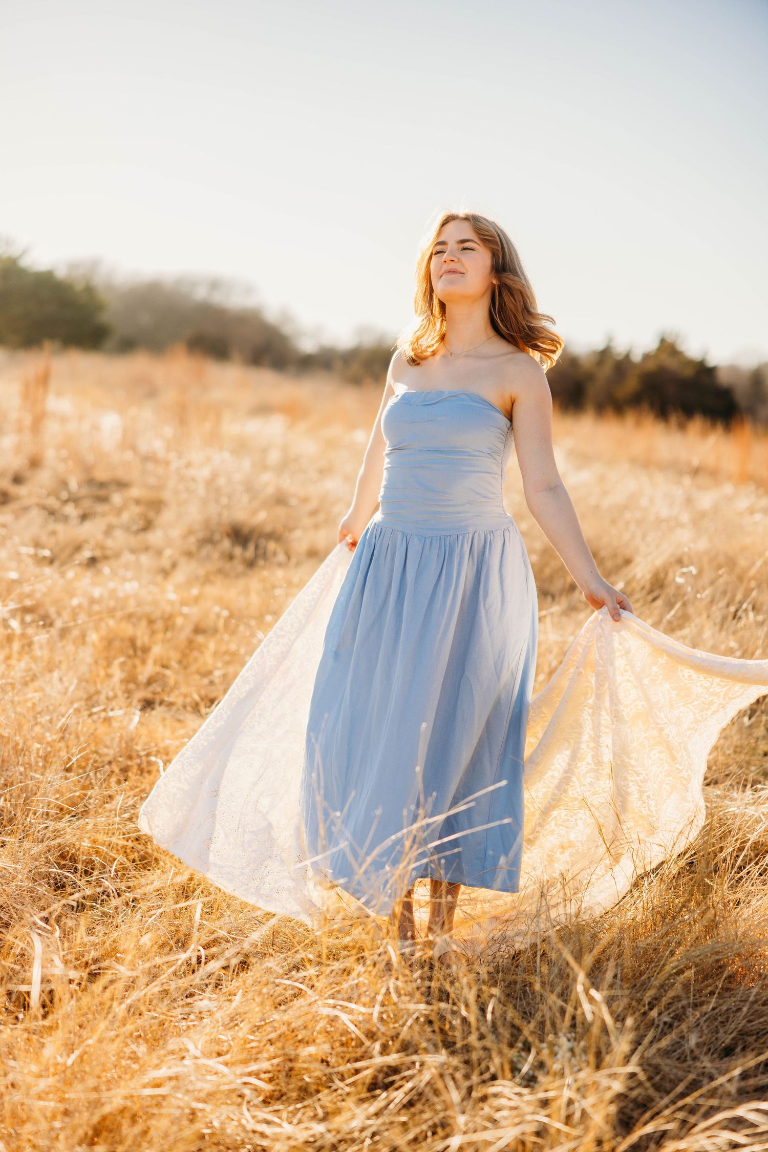 Young woman in a light blue strapless dress standing in a golden field, holding her skirt in warm sunlight.