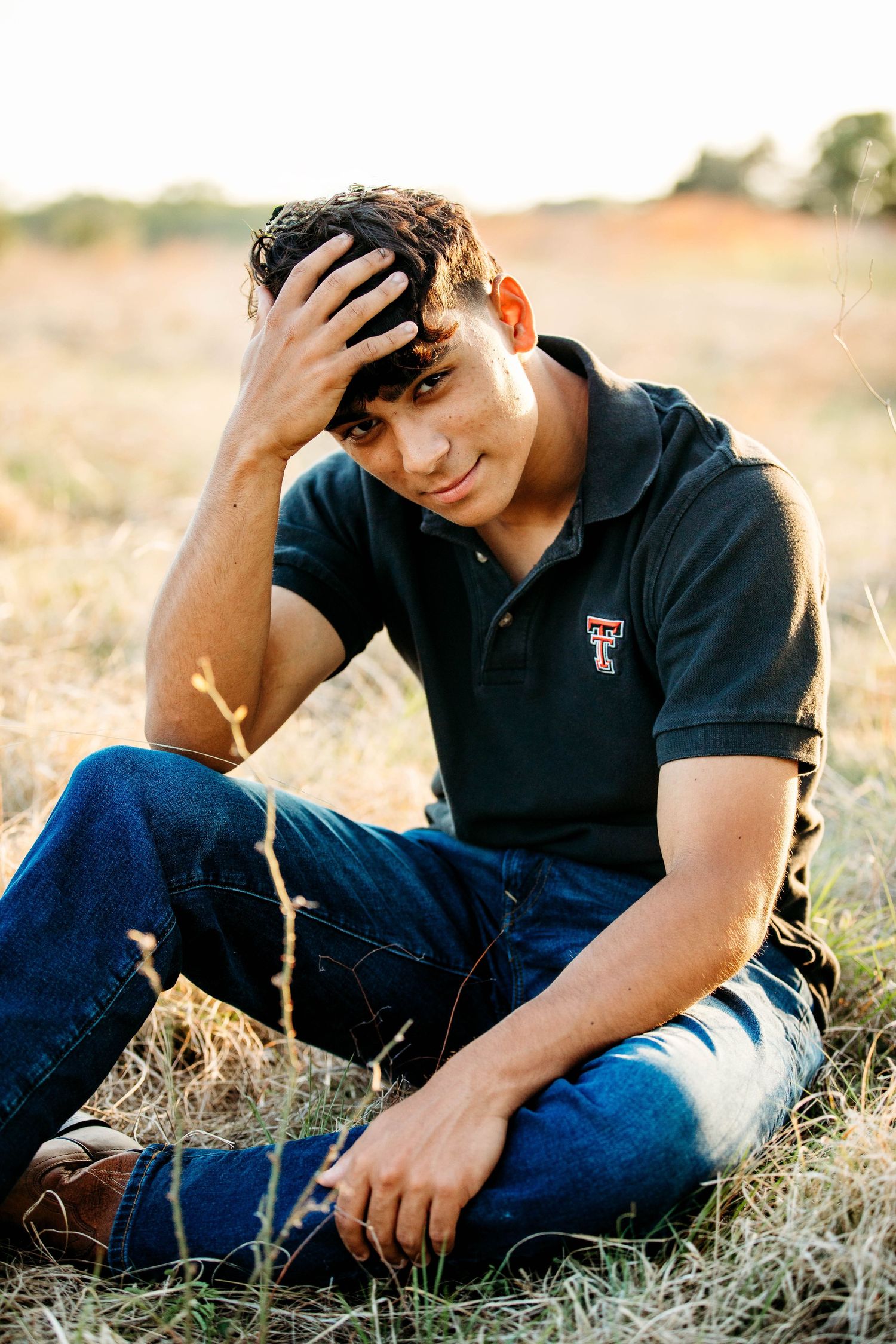 Young man in Texas Tech polo shirt sitting in a golden field, hand on head, wearing blue jeans.