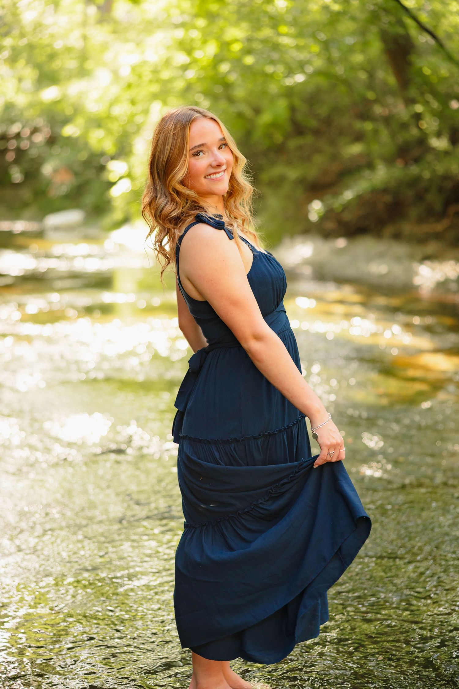 Young woman in navy blue dress smiling by a sunlit creek, surrounded by lush green trees.