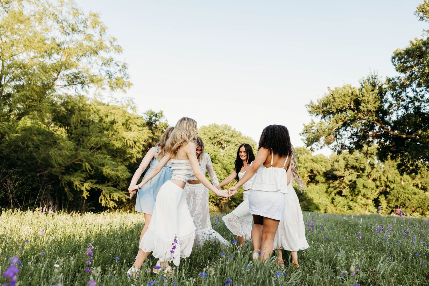 Four girls in white dresses hold hands dancing in a sunlit meadow with wildflowers and green trees.