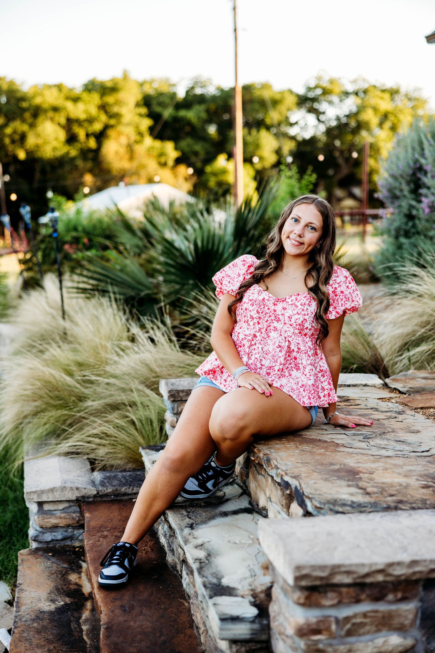 High school senior girl sitting on stone steps in a pink floral top and denim shorts, smiling during her outdoor portrait session.