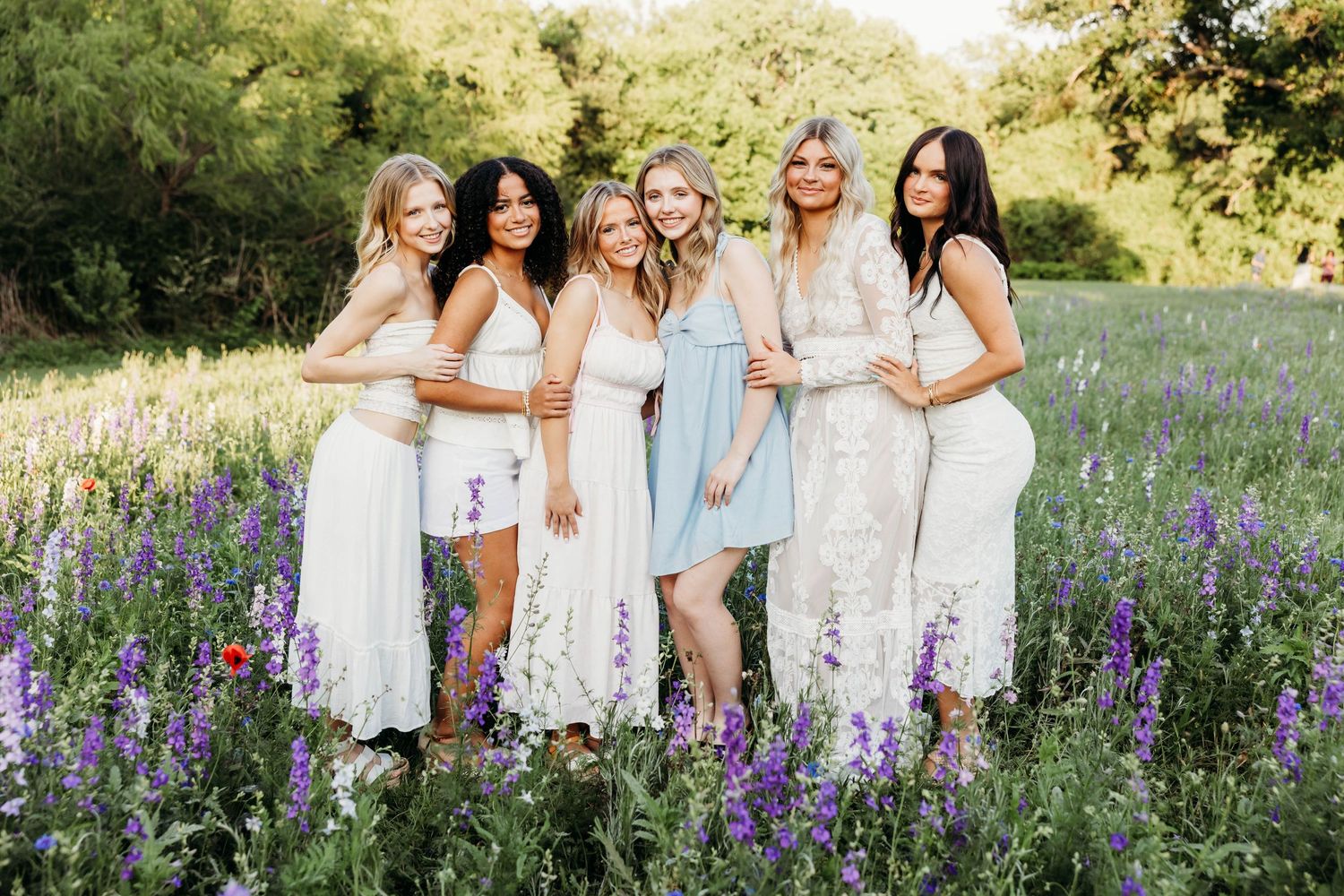 Six women in white and pastel dresses posing together in a vibrant wildflower field with purple blooms.