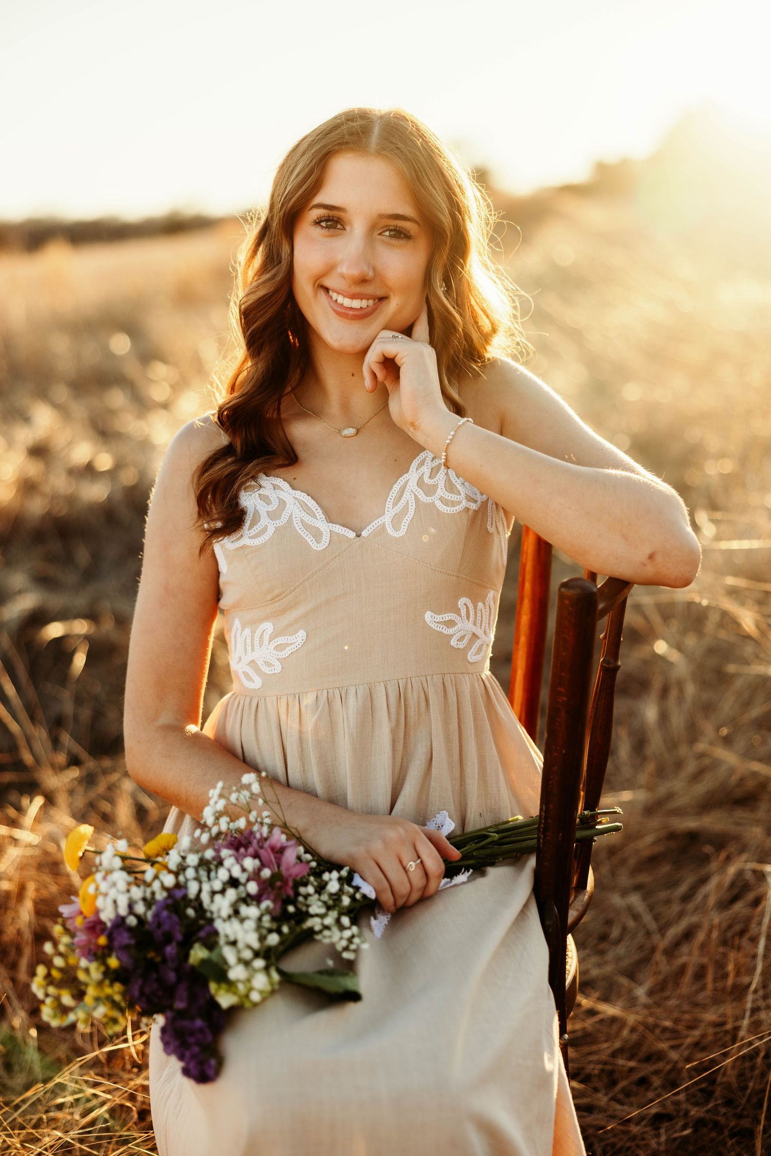 A person in a beige dress holds a bouquet of purple and white wildflowers during golden hour sunset.