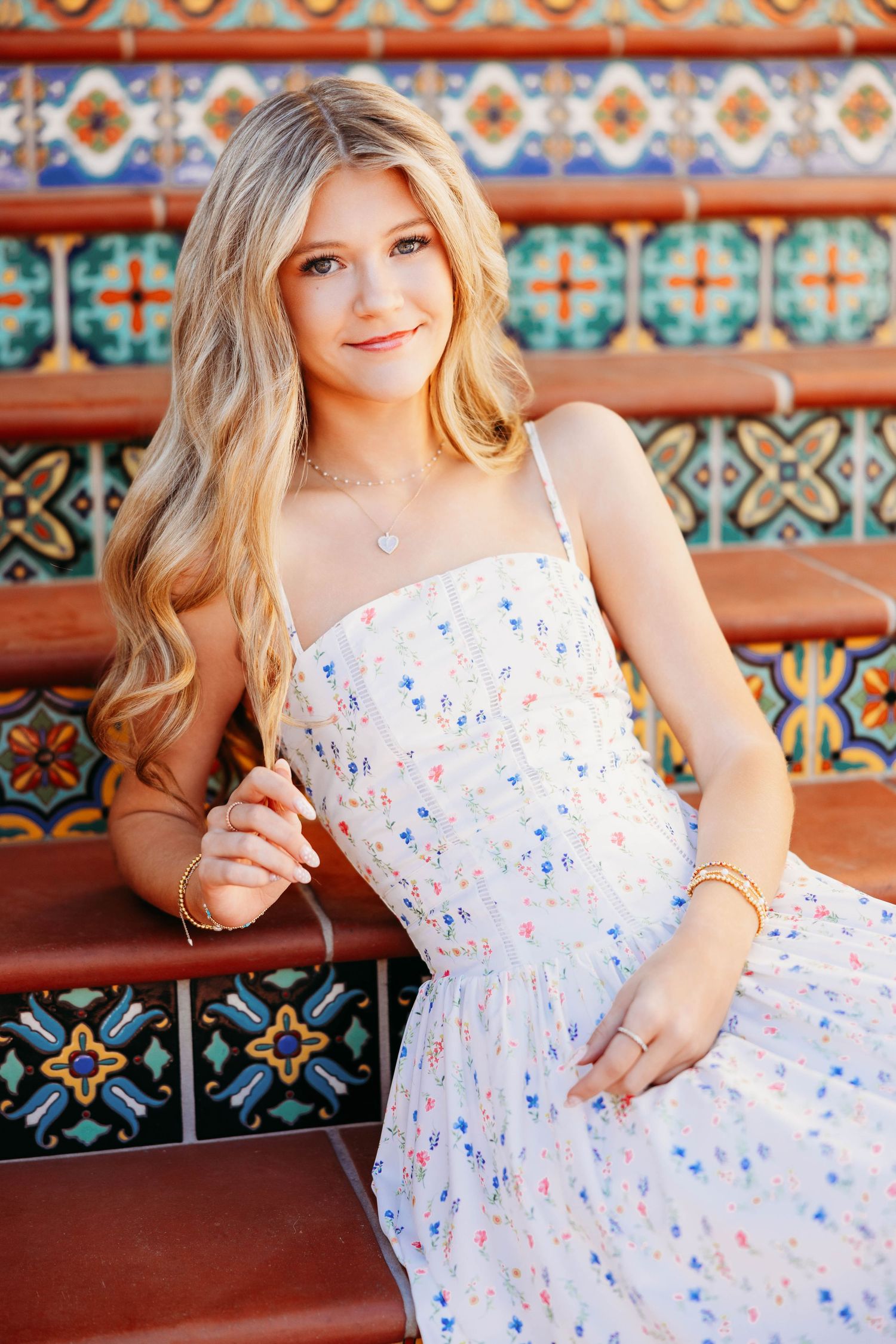 Young woman in floral sundress sitting on colorful tiled steps, smiling with long blonde hair.