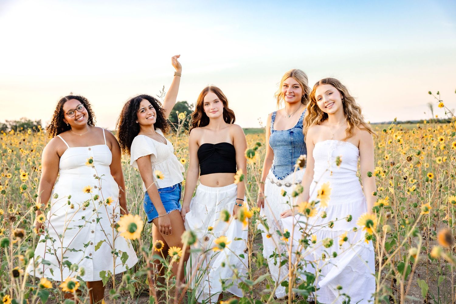 Friends in white dresses pose together in a blooming sunflower field at sunset.