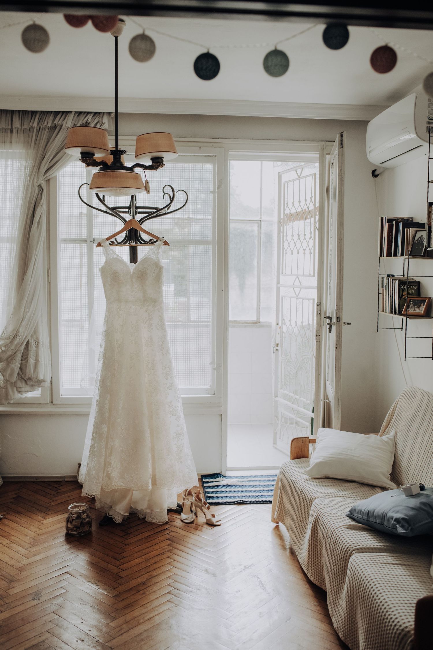 White lace wedding dress hanging in a bright, cozy room with herringbone wood floors and soft decor.