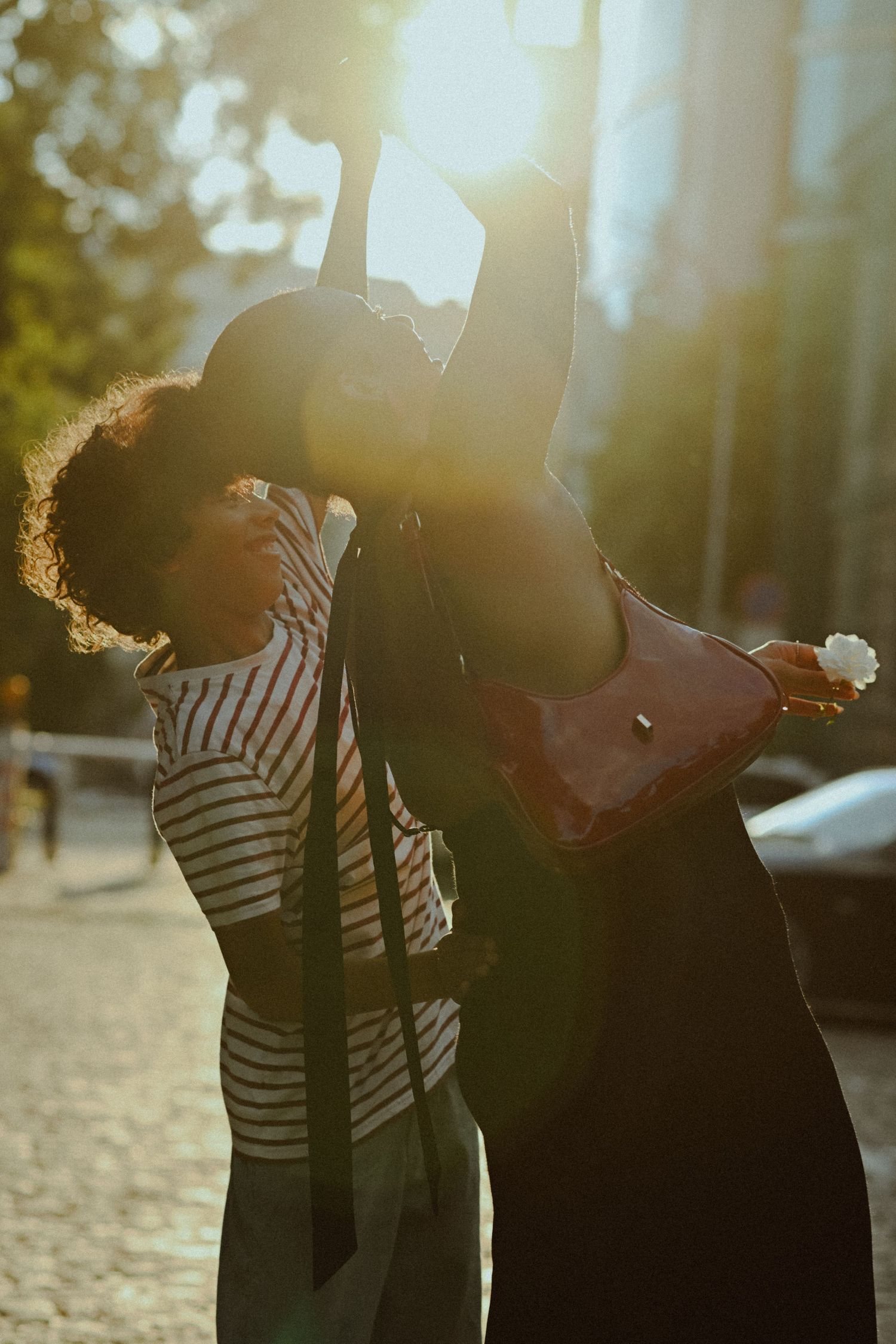 Two people dancing and posing together outdoors in backlit sunlight, one wearing stripes and one in dark clothing.