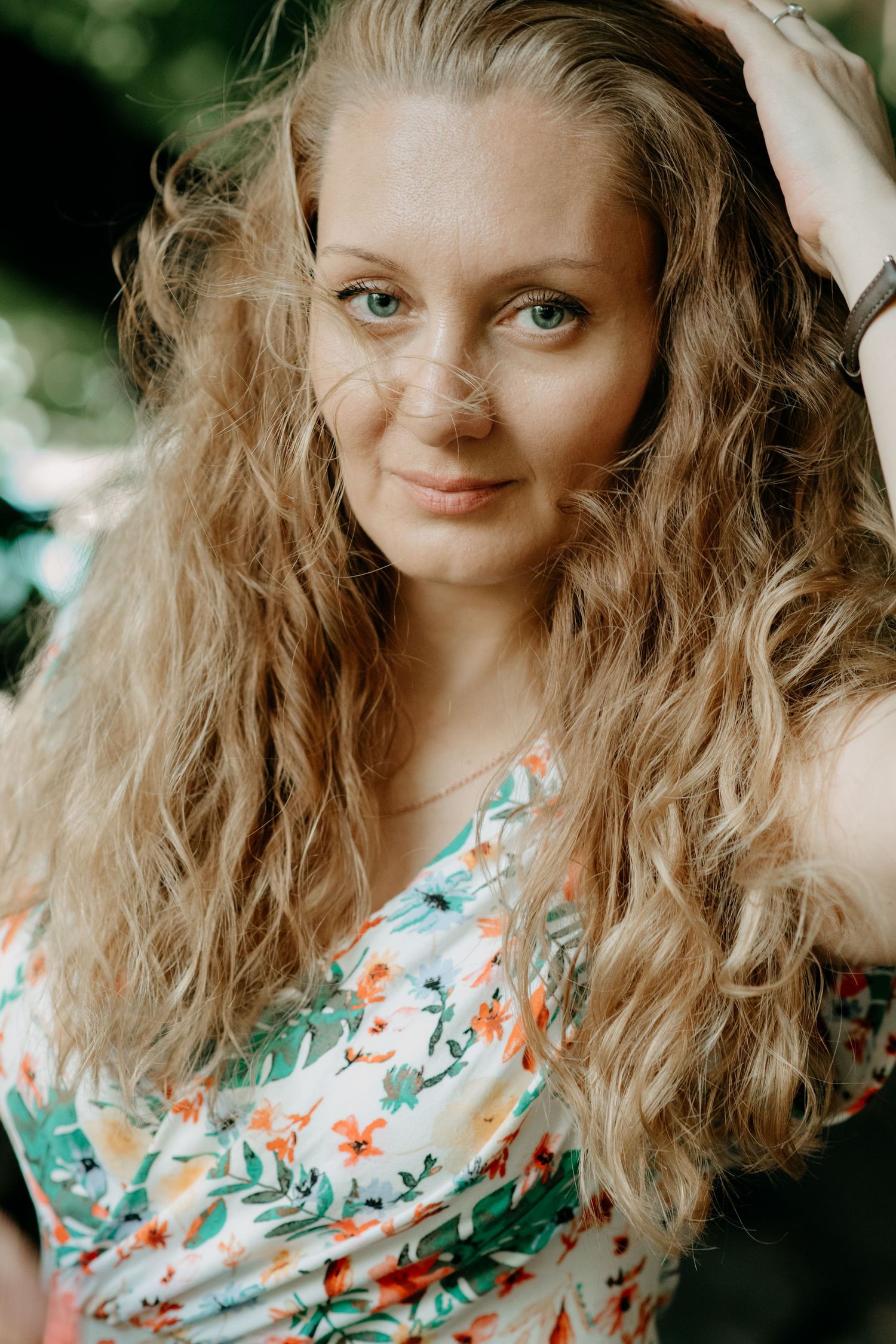 Young person with wavy blonde hair wearing a floral patterned dress in an outdoor setting.