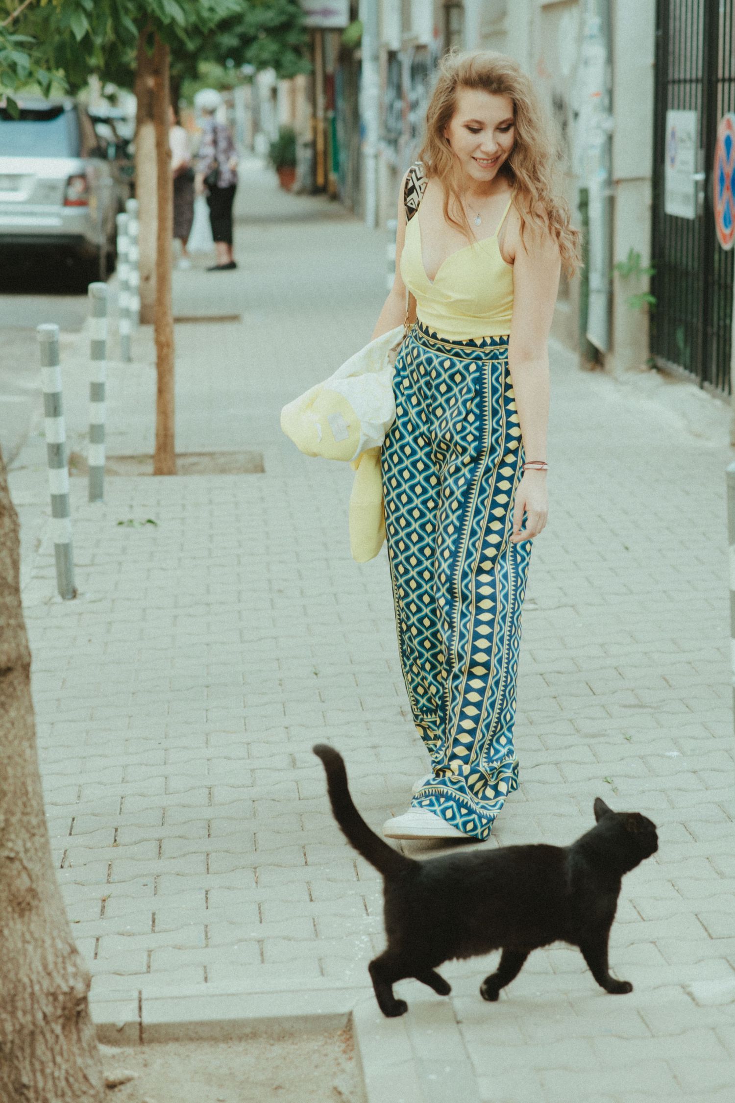 A candid street photo of someone in a yellow top and patterned pants walking past a black cat.