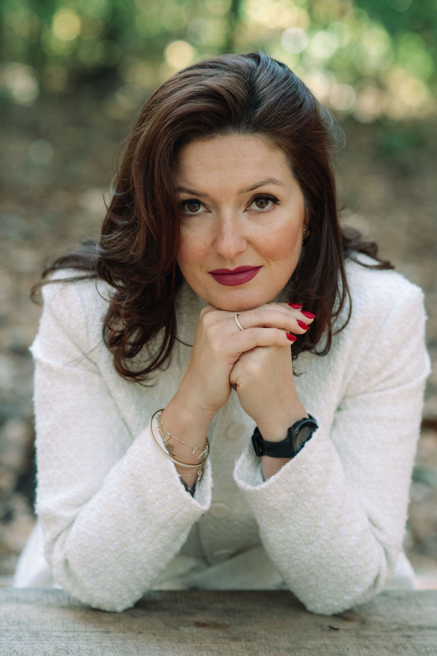 Portrait of someone in a white sweater with dark hair and red lipstick against a blurred natural background.