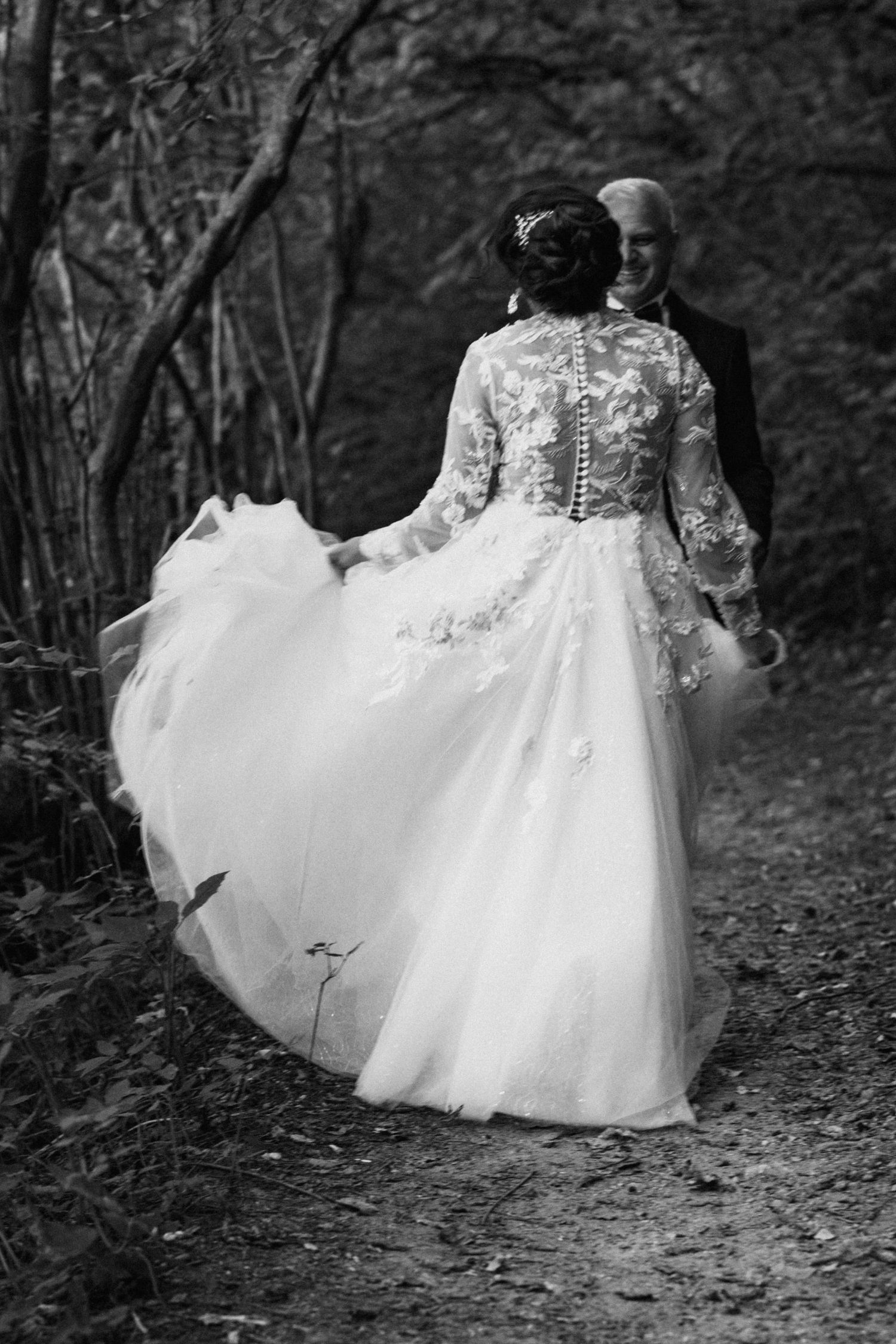 A flowing white wedding dress swirls dramatically on a forest path in black and white.