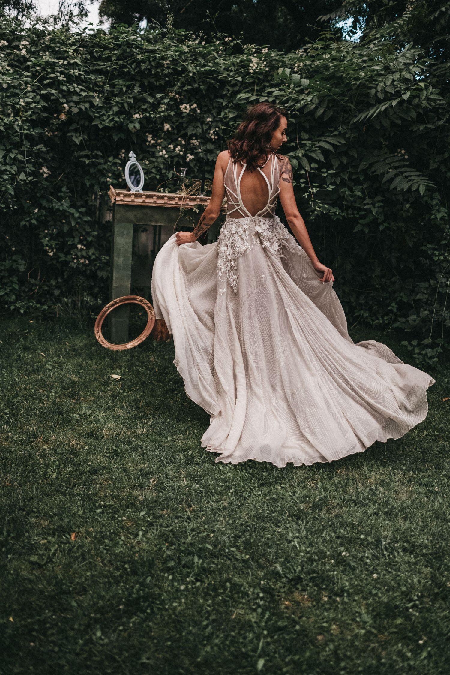 A flowing champagne-colored wedding dress with intricate floral details and open back design swirling on green grass.