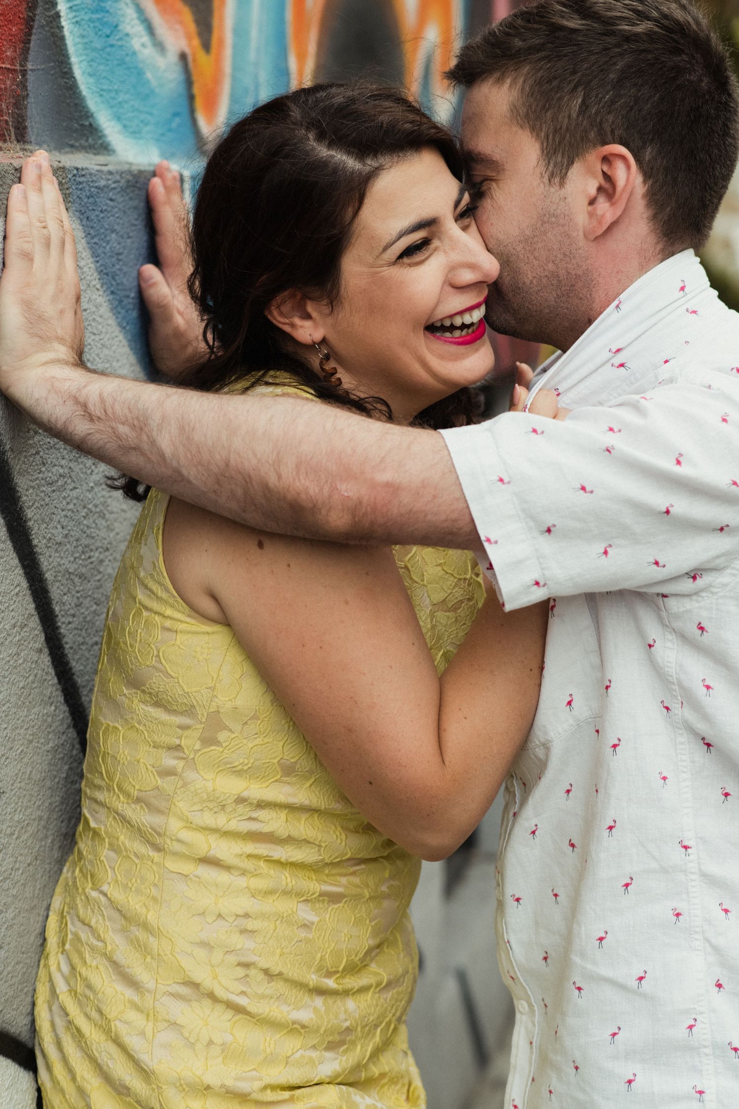 A laughing couple share an intimate moment against a colorful graffiti wall.