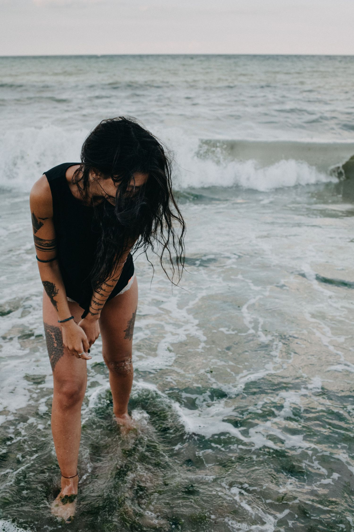 A person with dark hair stands in the ocean waves wearing a black swimsuit at the beach.