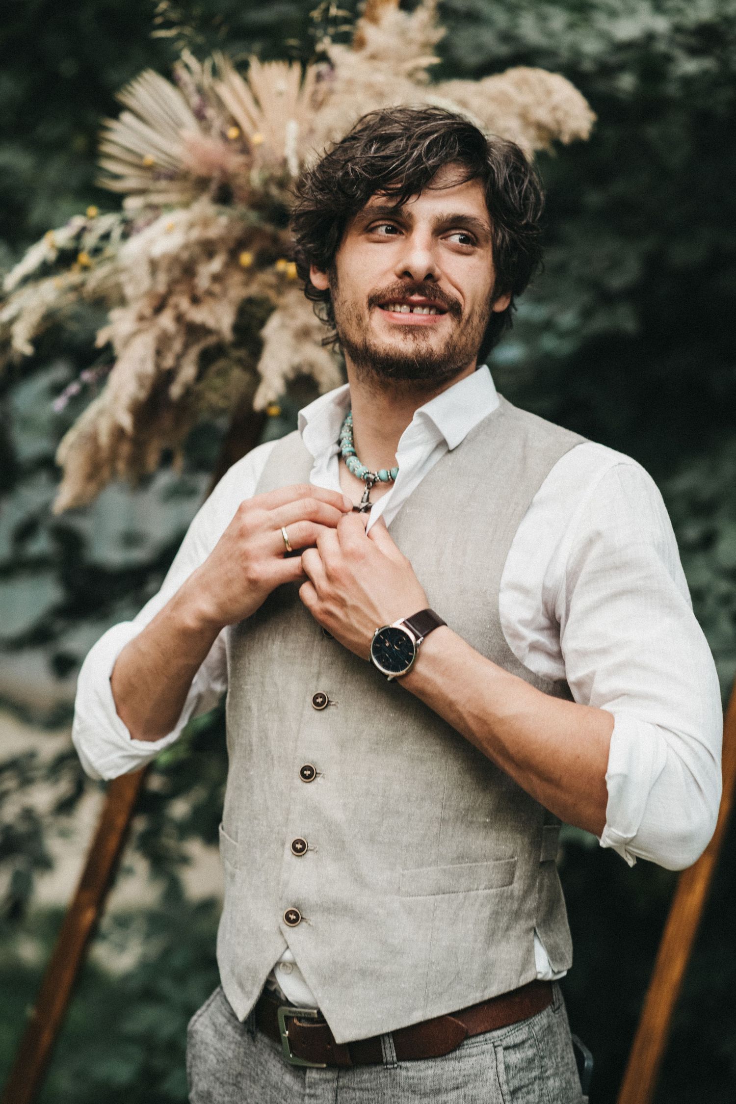 Stylish groom in beige vest and white shirt poses against natural outdoor backdrop.