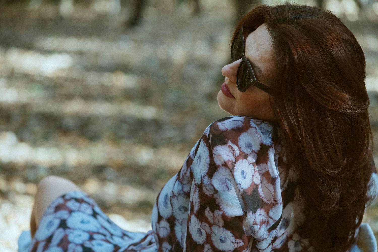 Person in floral dress and sunglasses relaxing outdoors in natural lighting.