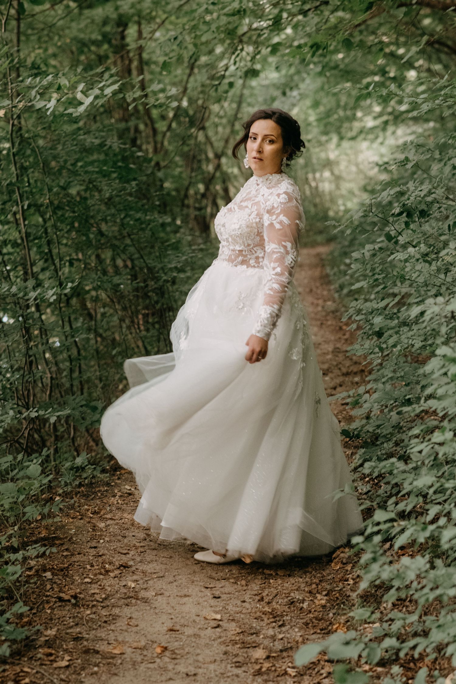 A bride in a long-sleeved lace wedding gown poses on a forest trail.