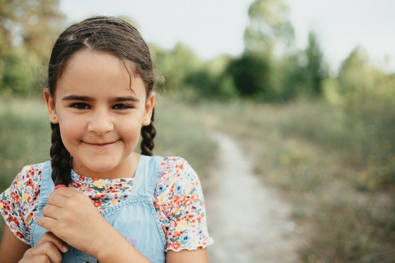 A child smiles warmly on a rural path wearing floral print and denim overalls.