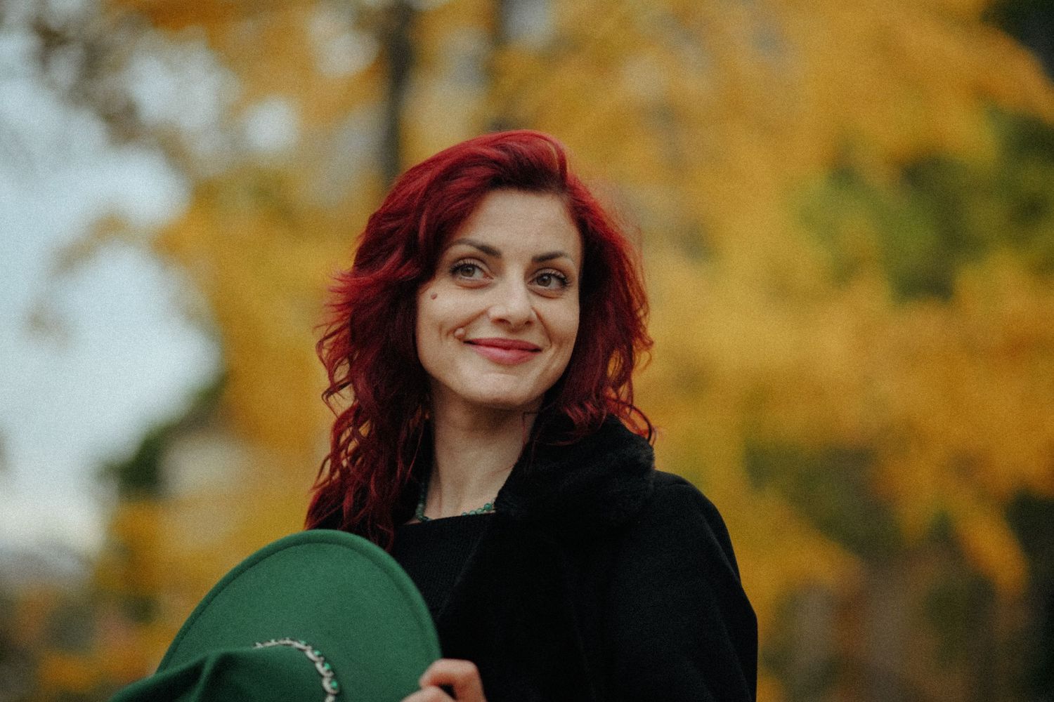 Woman with red hair poses outdoors against a vibrant autumn background with golden foliage.