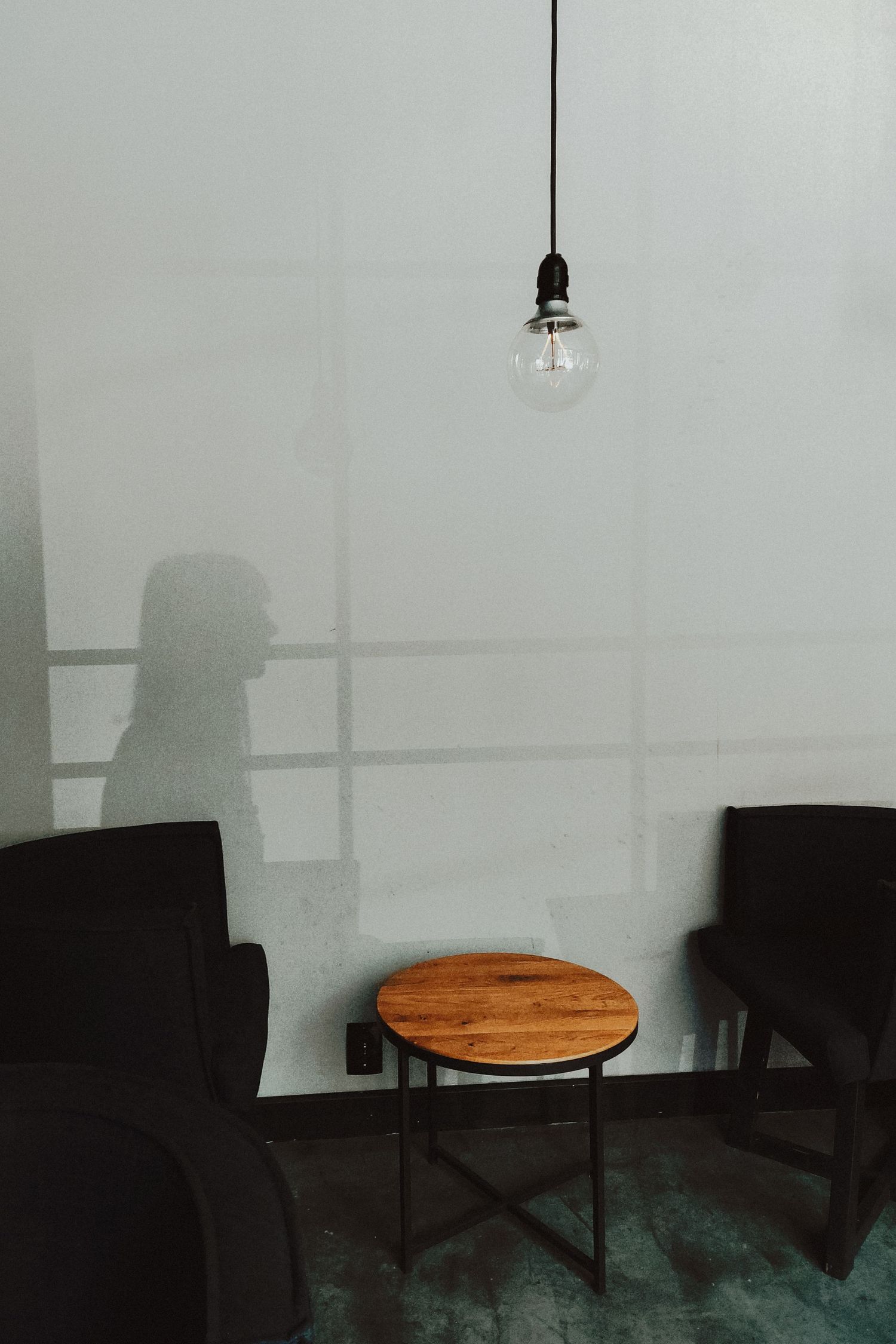 Minimalist room interior with wooden side table, black chairs and hanging light bulb casting shadows.