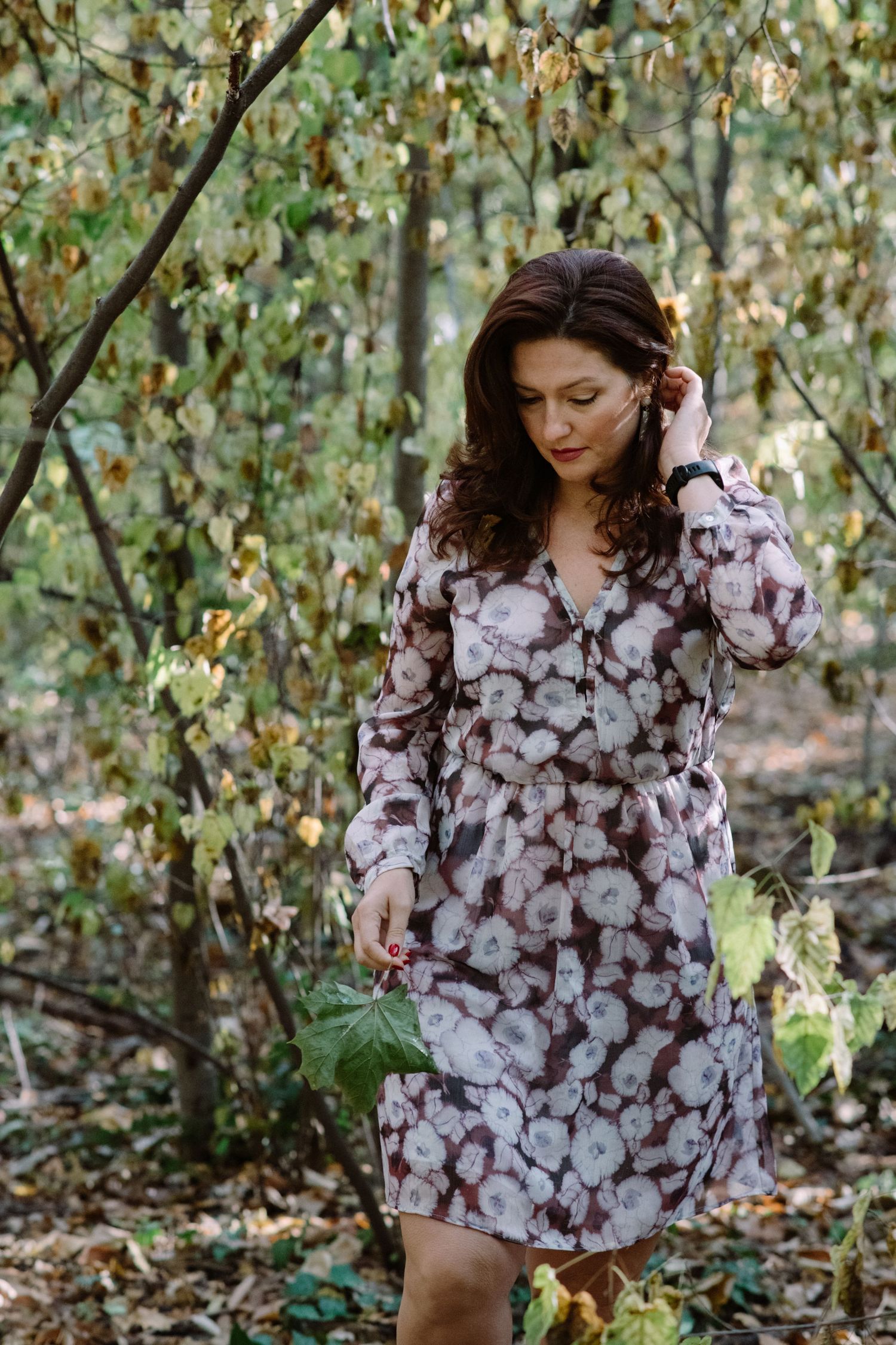Woman in a floral print dress walking through a wooded area with autumn foliage.