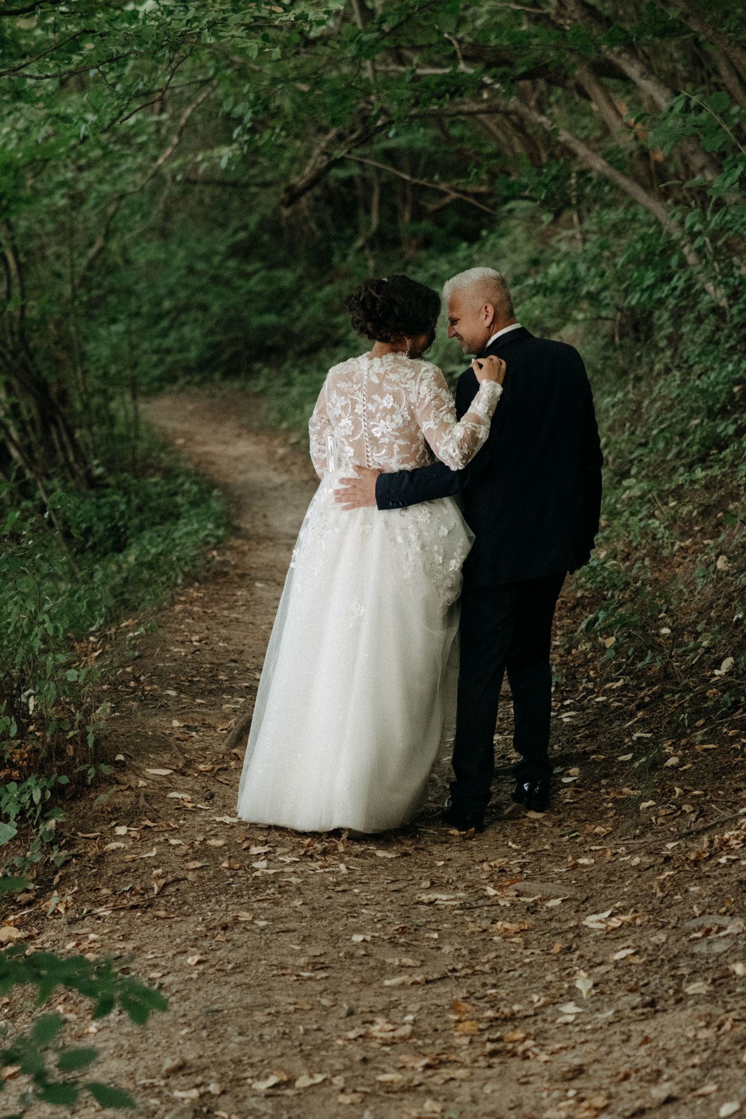 A romantic wedding couple walks down a wooded path in elegant attire.
