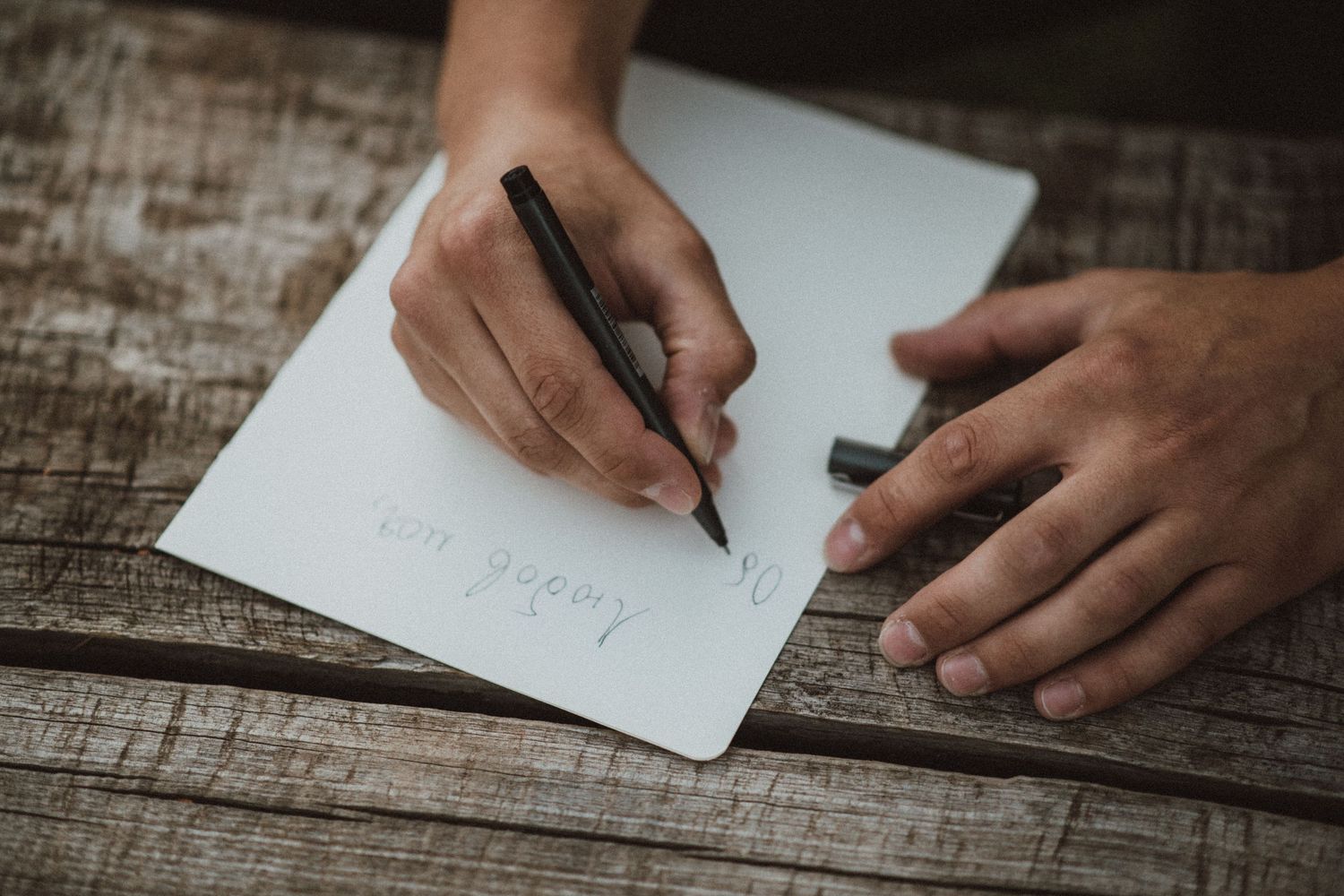 Hands writing with a pen on white paper at a wooden table.