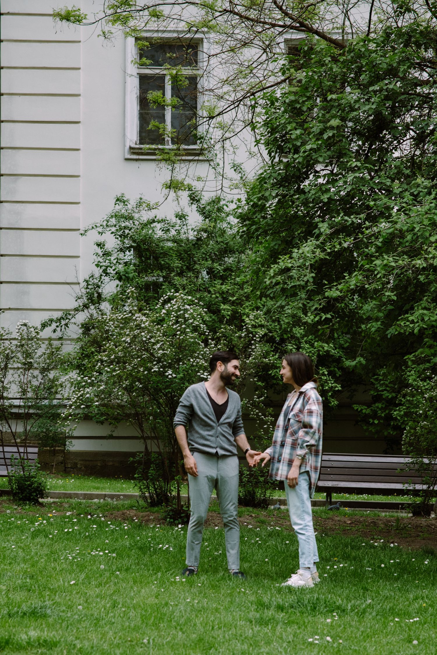 Couple holding hands while standing in front of a white building surrounded by greenery.