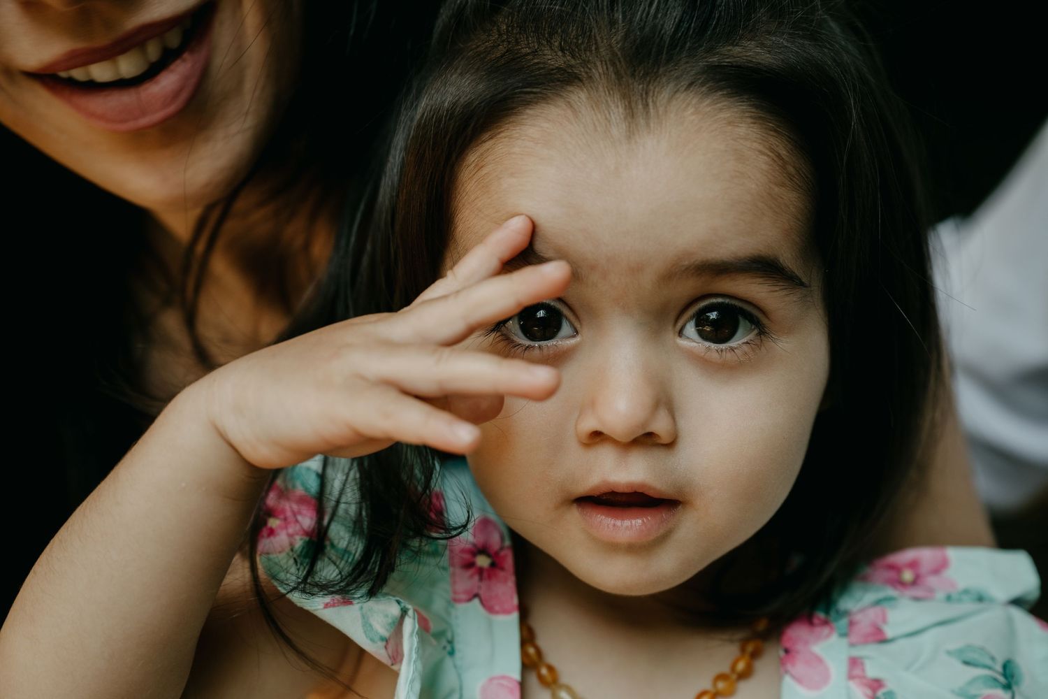 Young child in floral print clothing holds hand near face while looking to the side with wide-eyed expression.