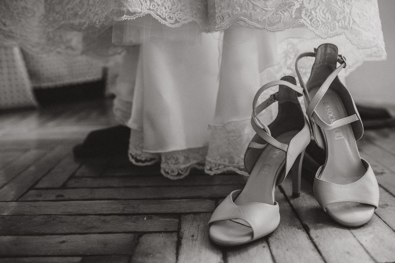 Elegant white bridal shoes next to the hem of a lace wedding dress on wooden flooring.