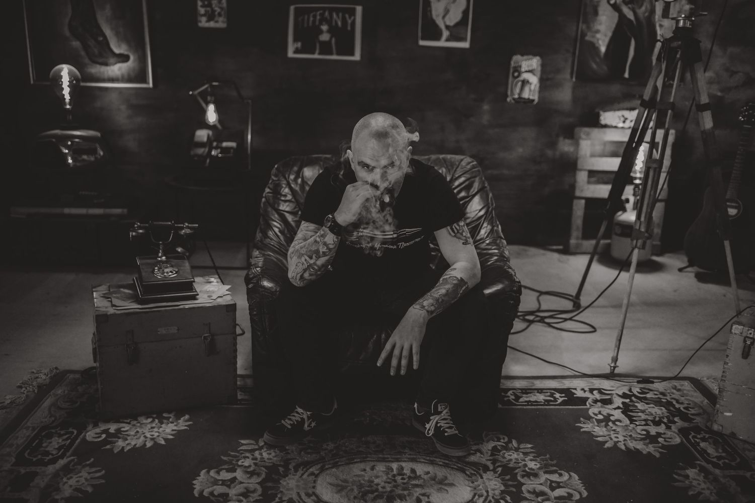 A moody black and white portrait shows a person with tattoos sitting in a dimly lit room with a patterned rug.