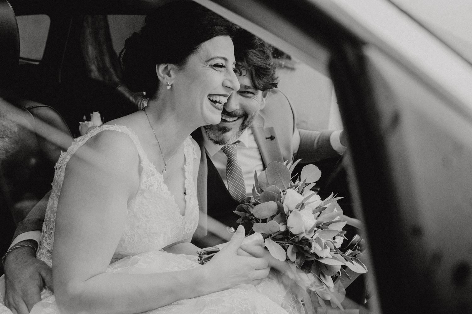 Black and white candid wedding photo capturing joyful laughter in a car.