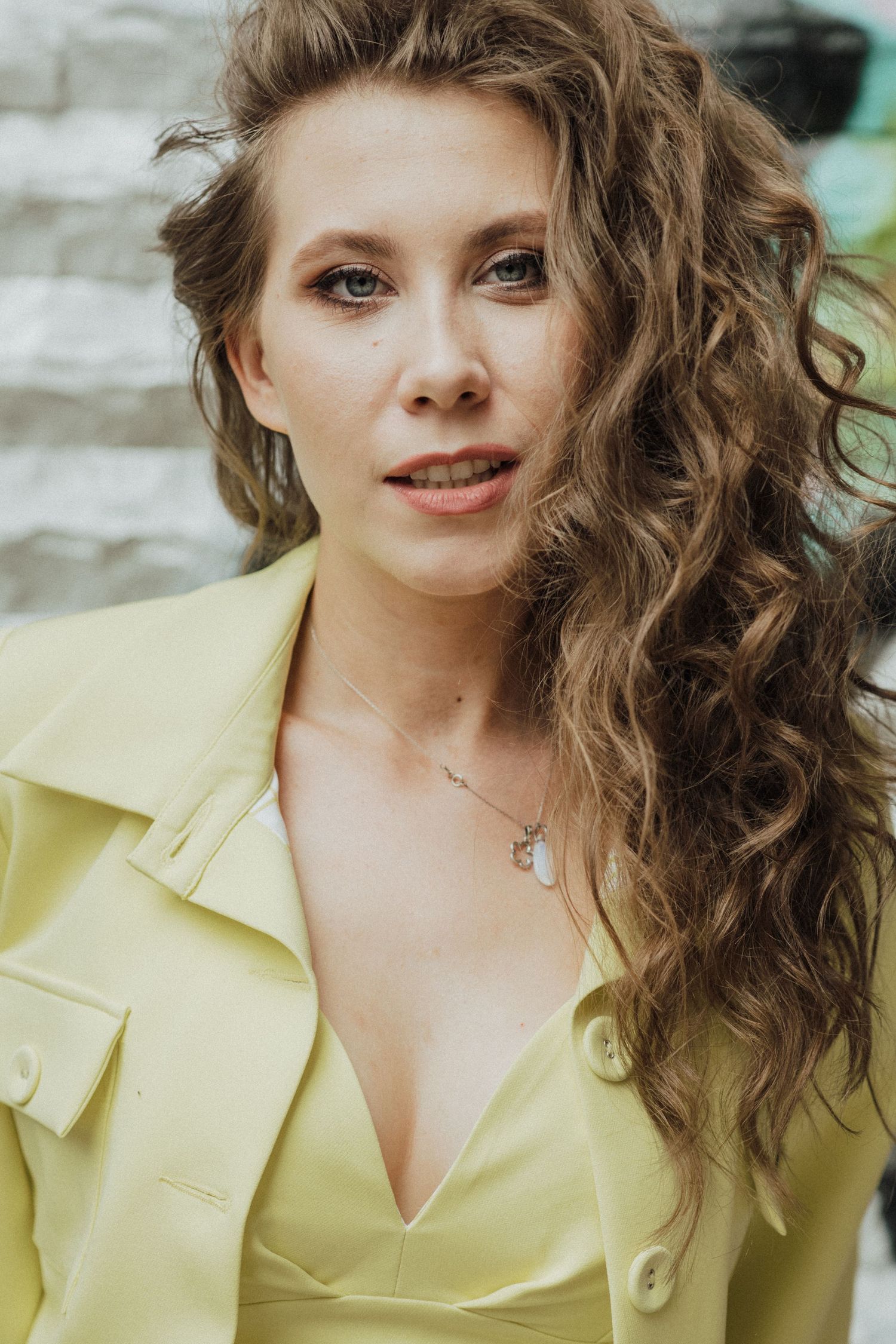 A portrait outdoors featuring someone in a pale yellow blazer with flowing curly hair.