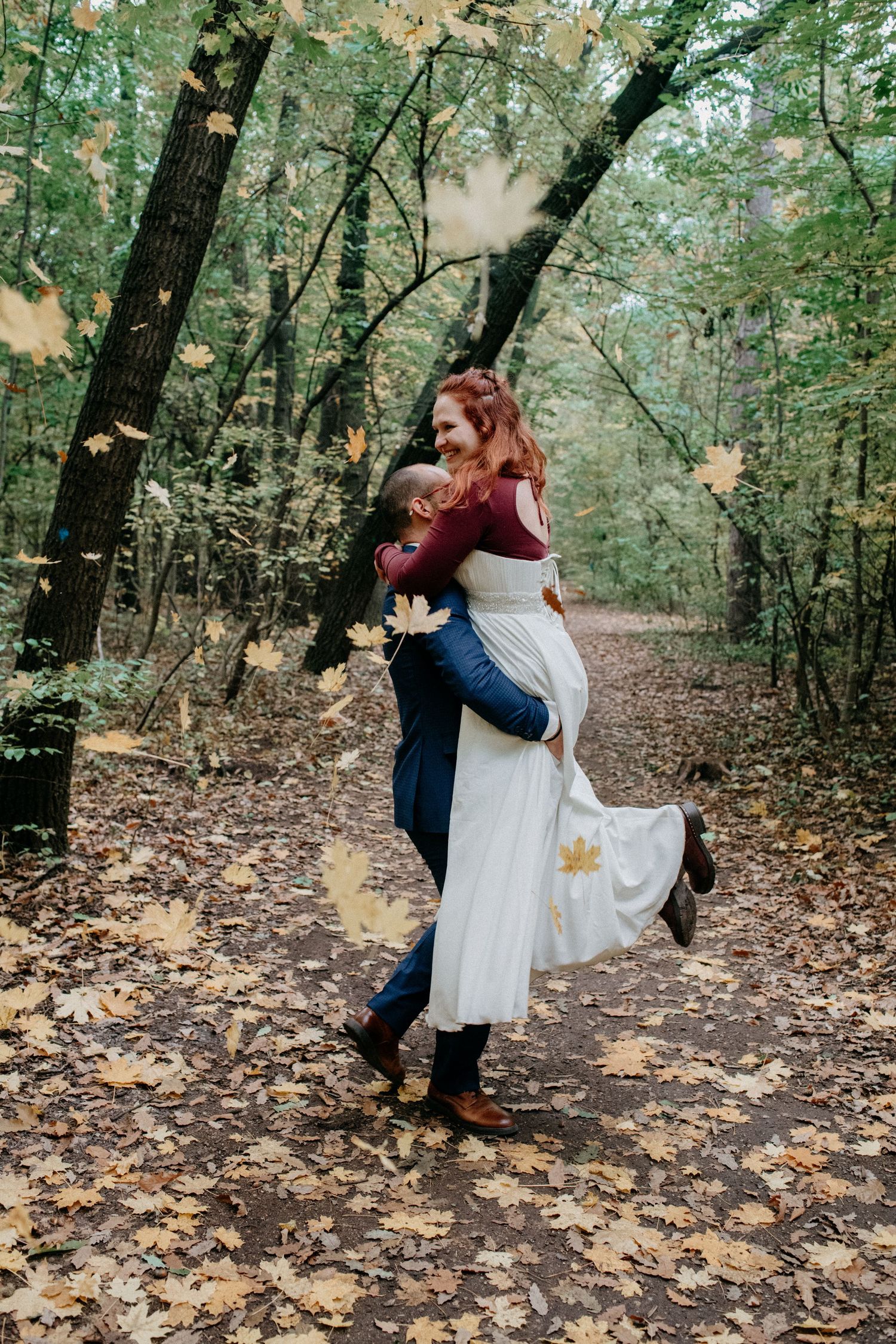 A couple embraces in an autumn forest with falling leaves and a flowing white dress.