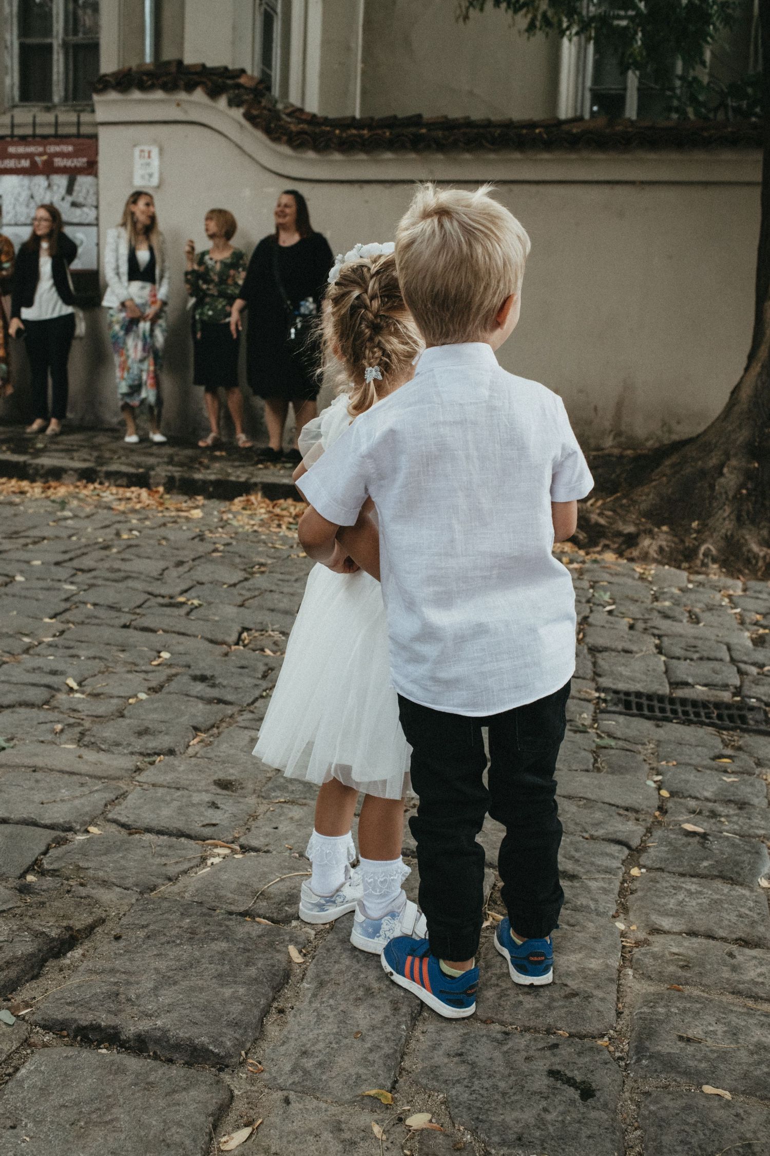Two small children walking together on a cobblestone path wearing formal attire.