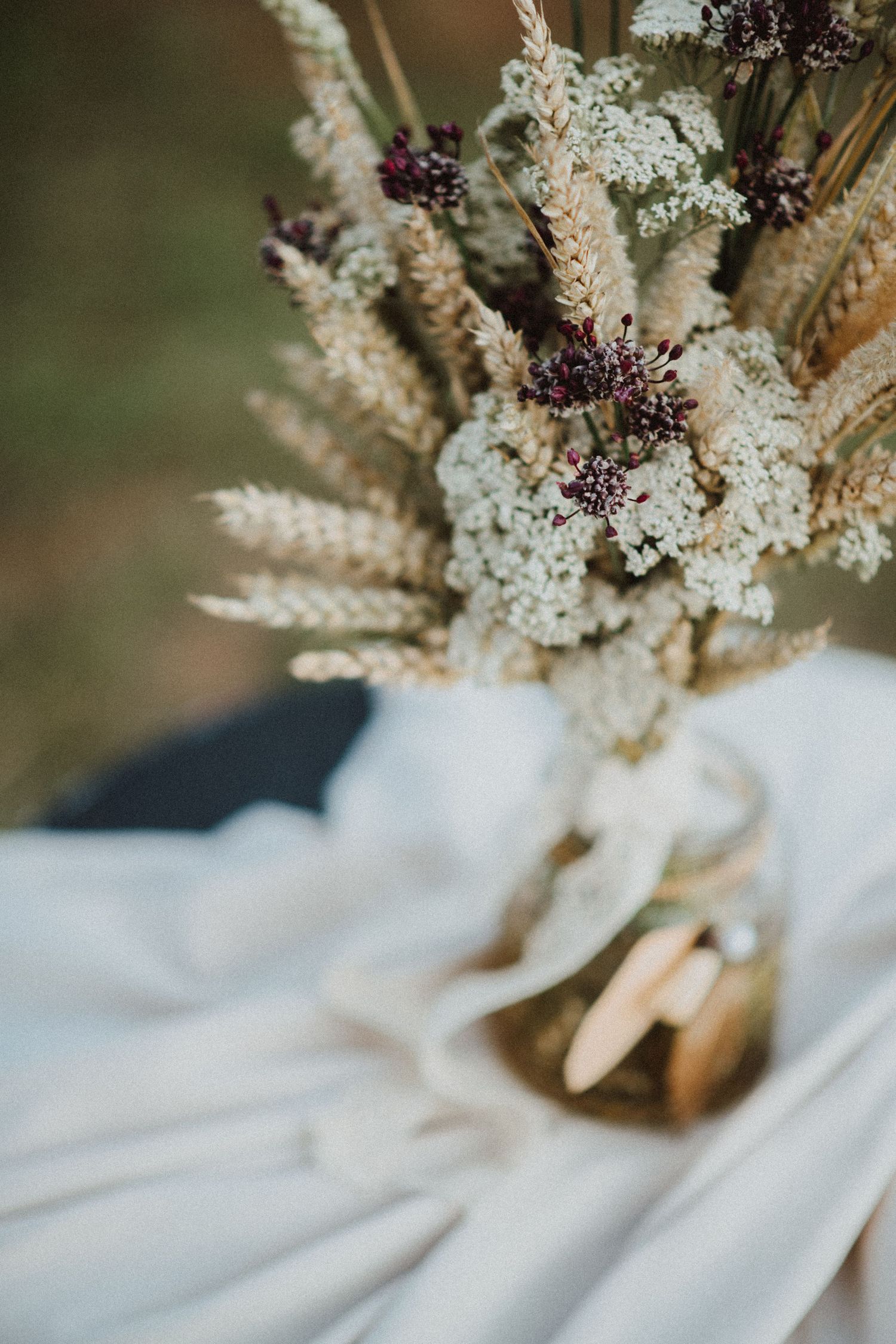Rustic bouquet of dried wheat stalks and delicate white flowers arranged in a golden vase on white fabric.