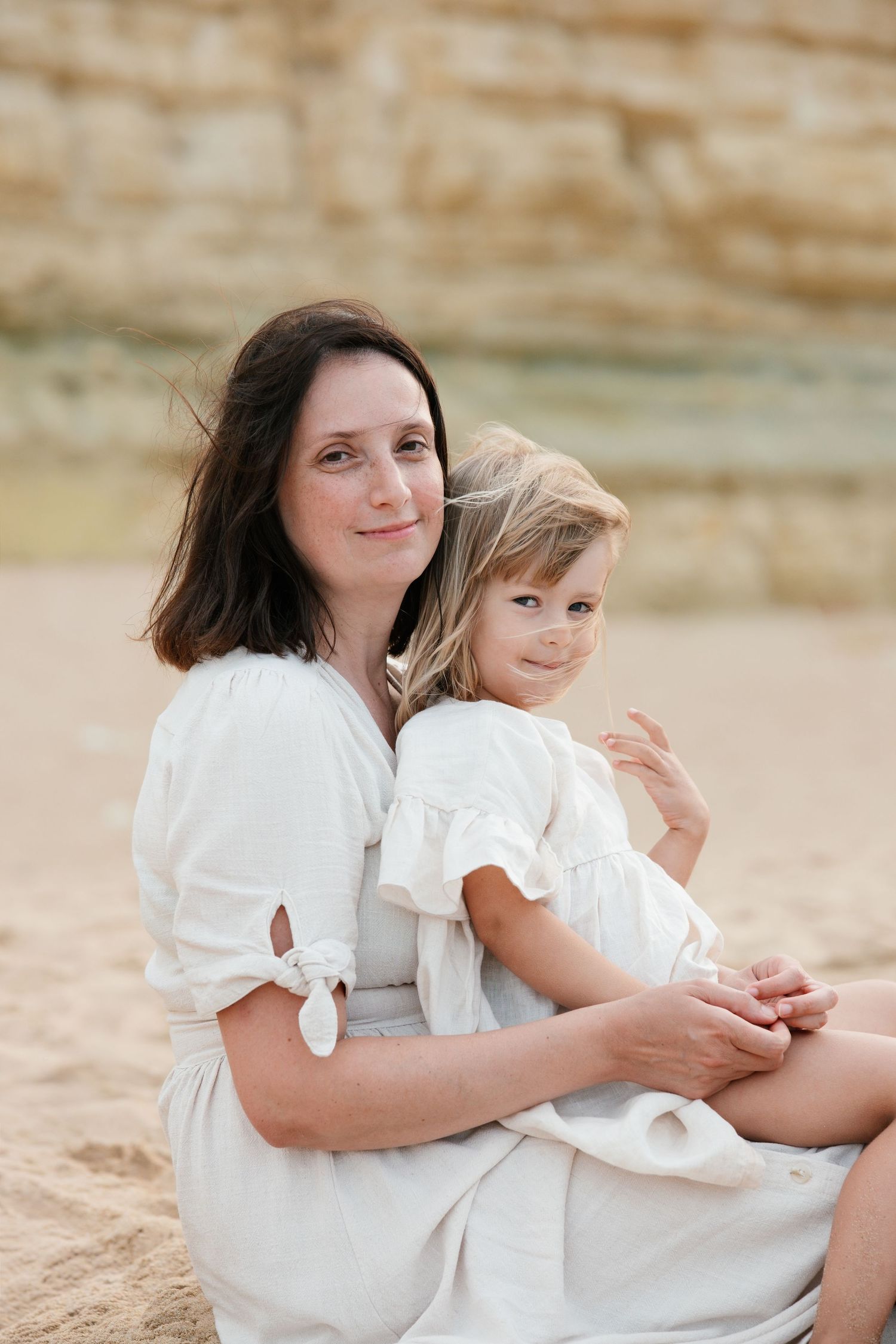 Mother and daughter cuddling on sandy shoreline with soft morning light in Lagos, Portugal.
