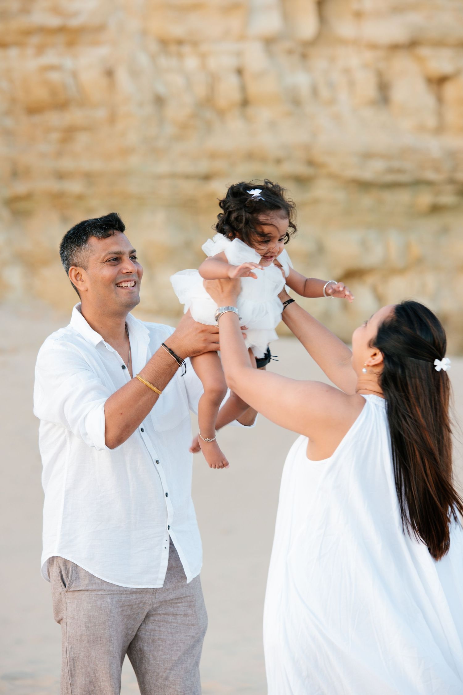 baby smiling while being lifted in the air by parents at Porto de Mós beach with cliffs in background