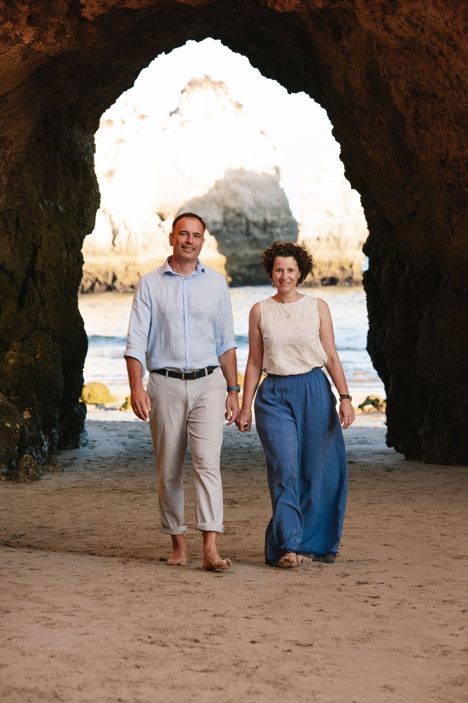candid moment of parents walking under the iconic arch at Praia Três Irmãos with ocean behind