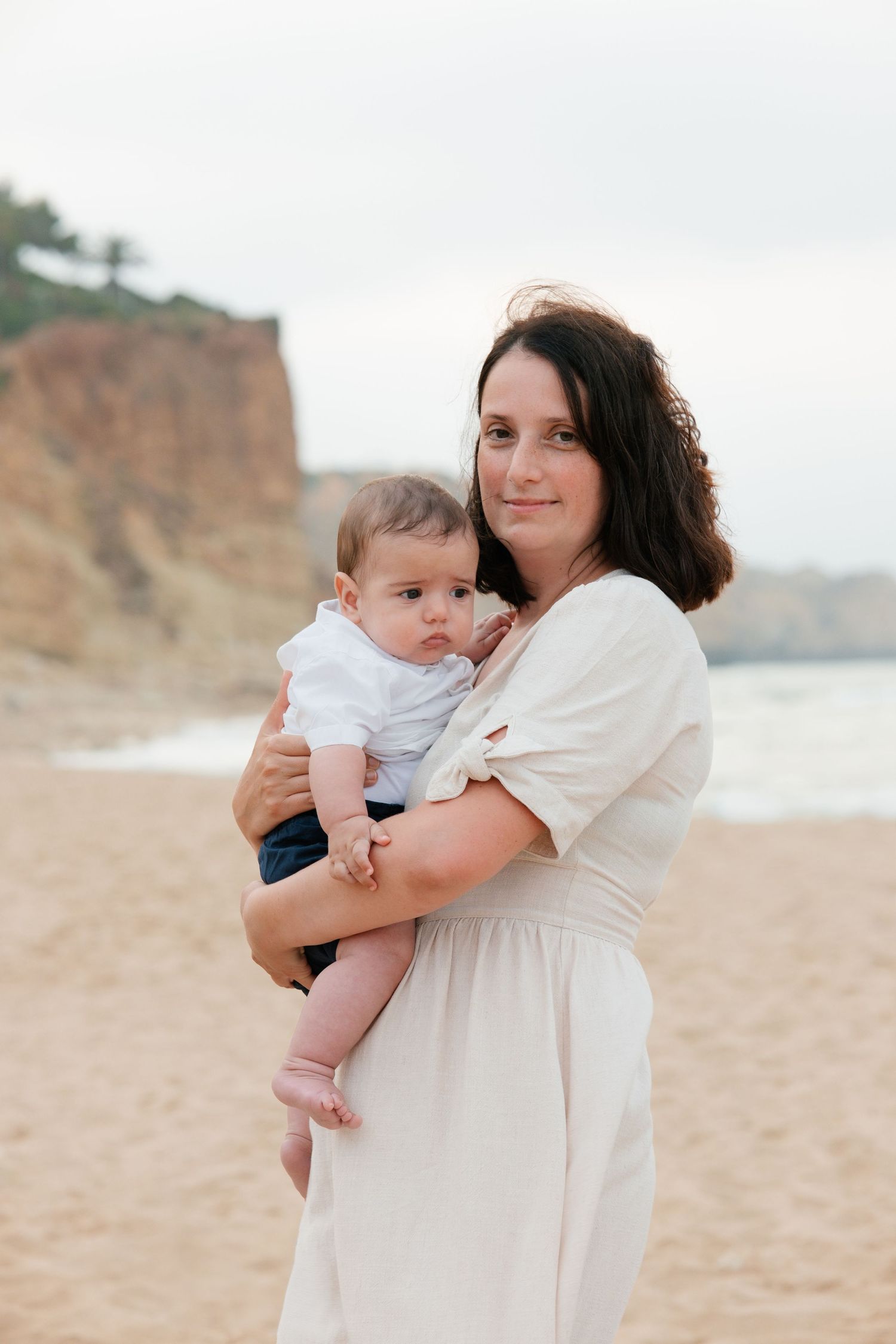 Mother cuddling baby close on sandy shoreline with soft morning light in Lagos, Portugal.