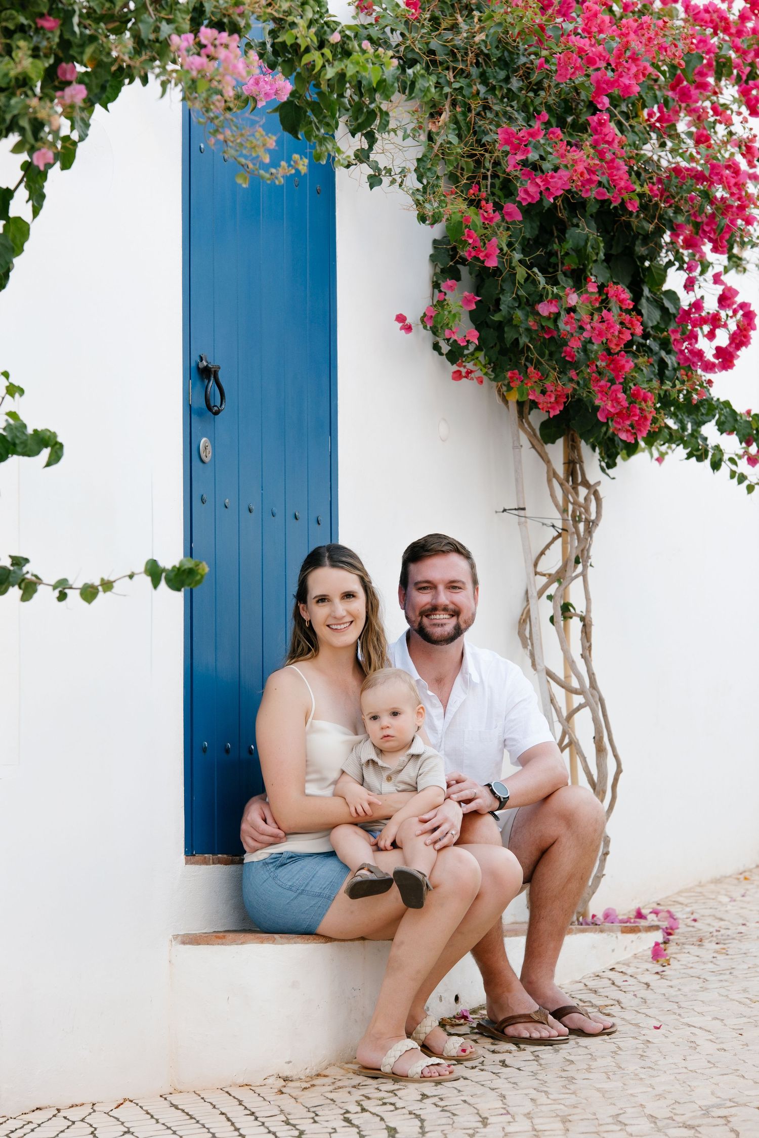 parents holding toddler while sitting on stone steps next to blue Algarve door and floral vines