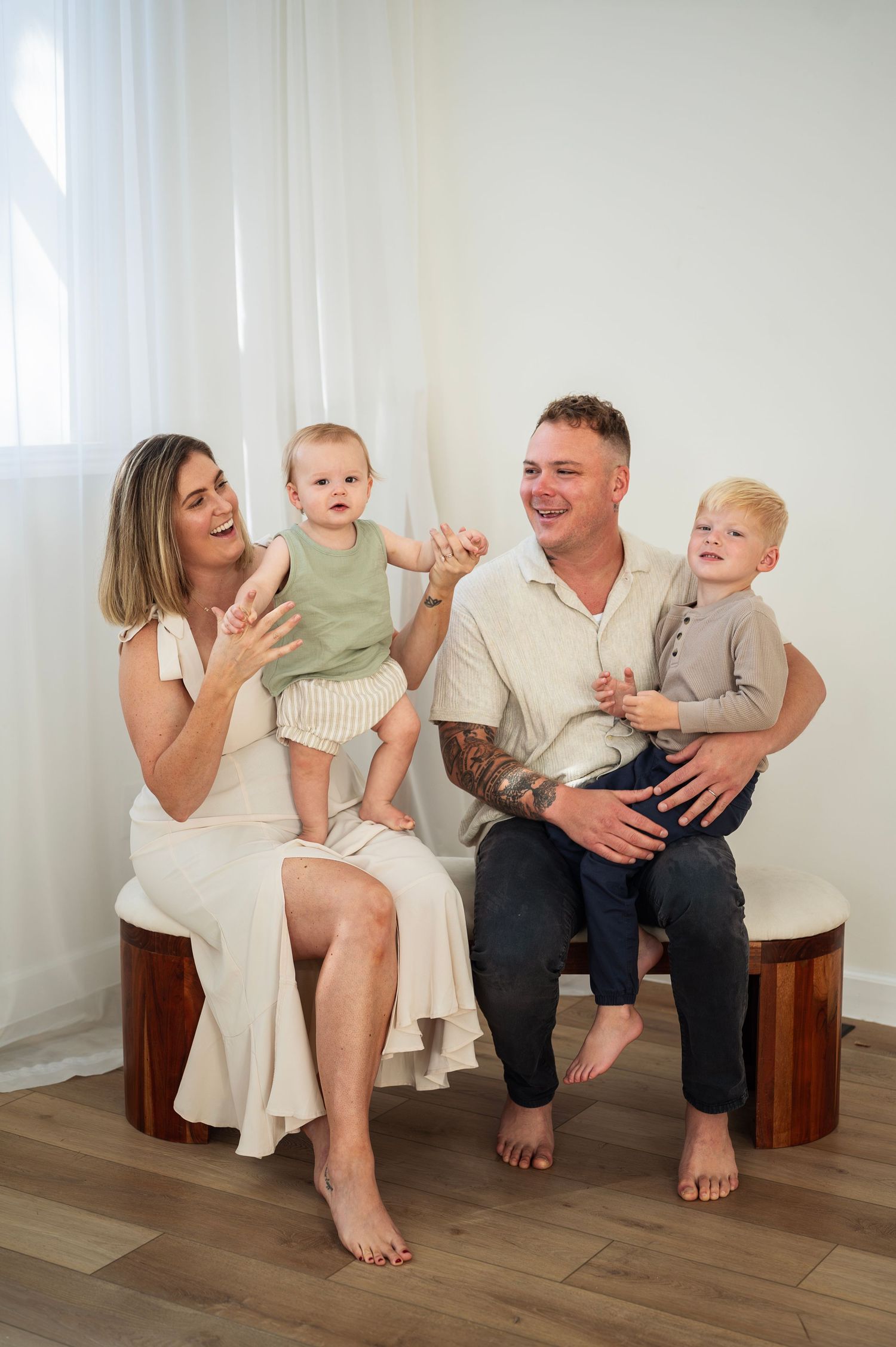 Family of four laughing together in a bright studio, parents holding two young children while sitting barefoot.