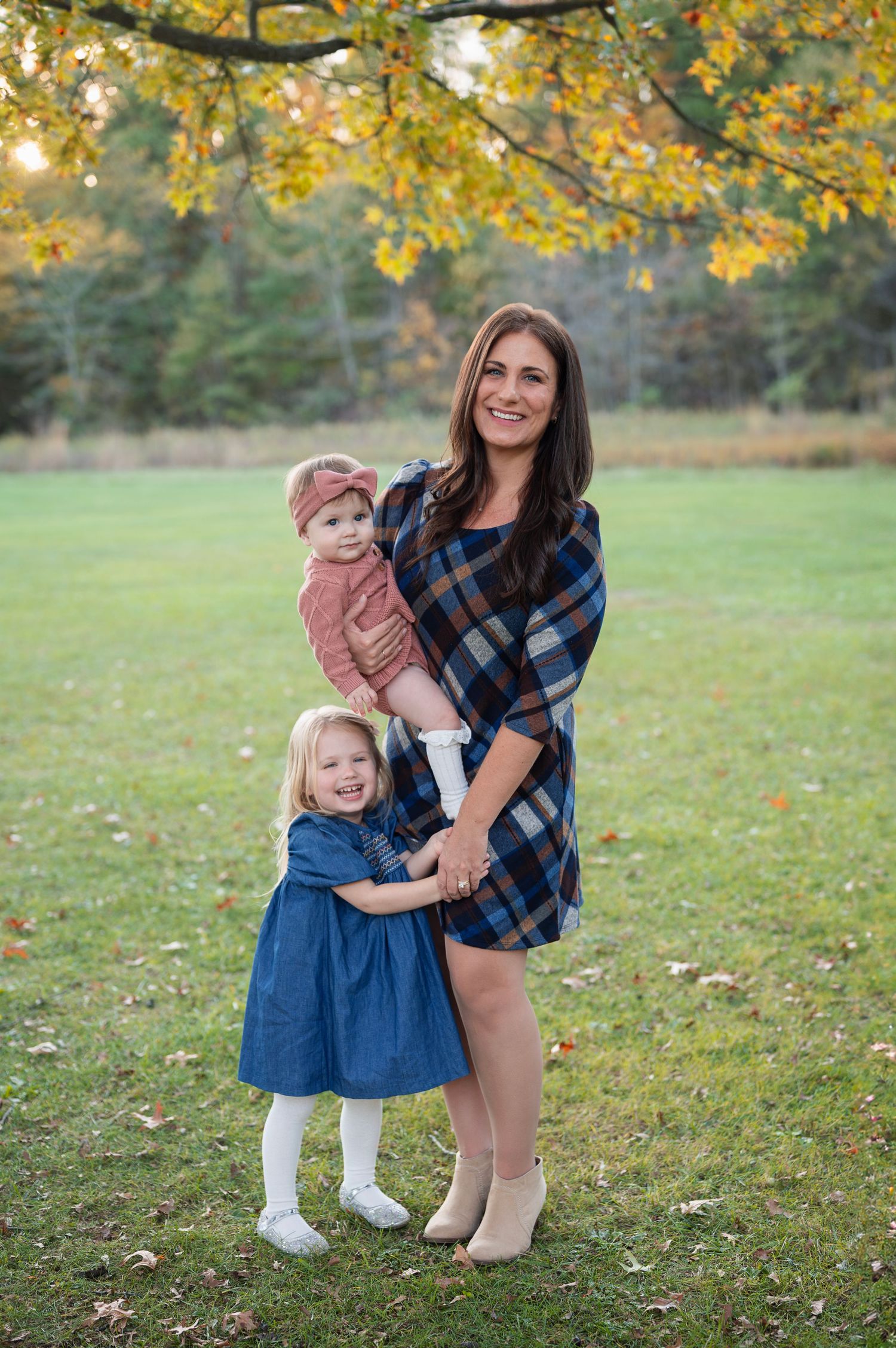 Mother in a plaid dress holding her baby and hugging her older daughter, both girls wearing coordinating pink and blue dresses, under a tree