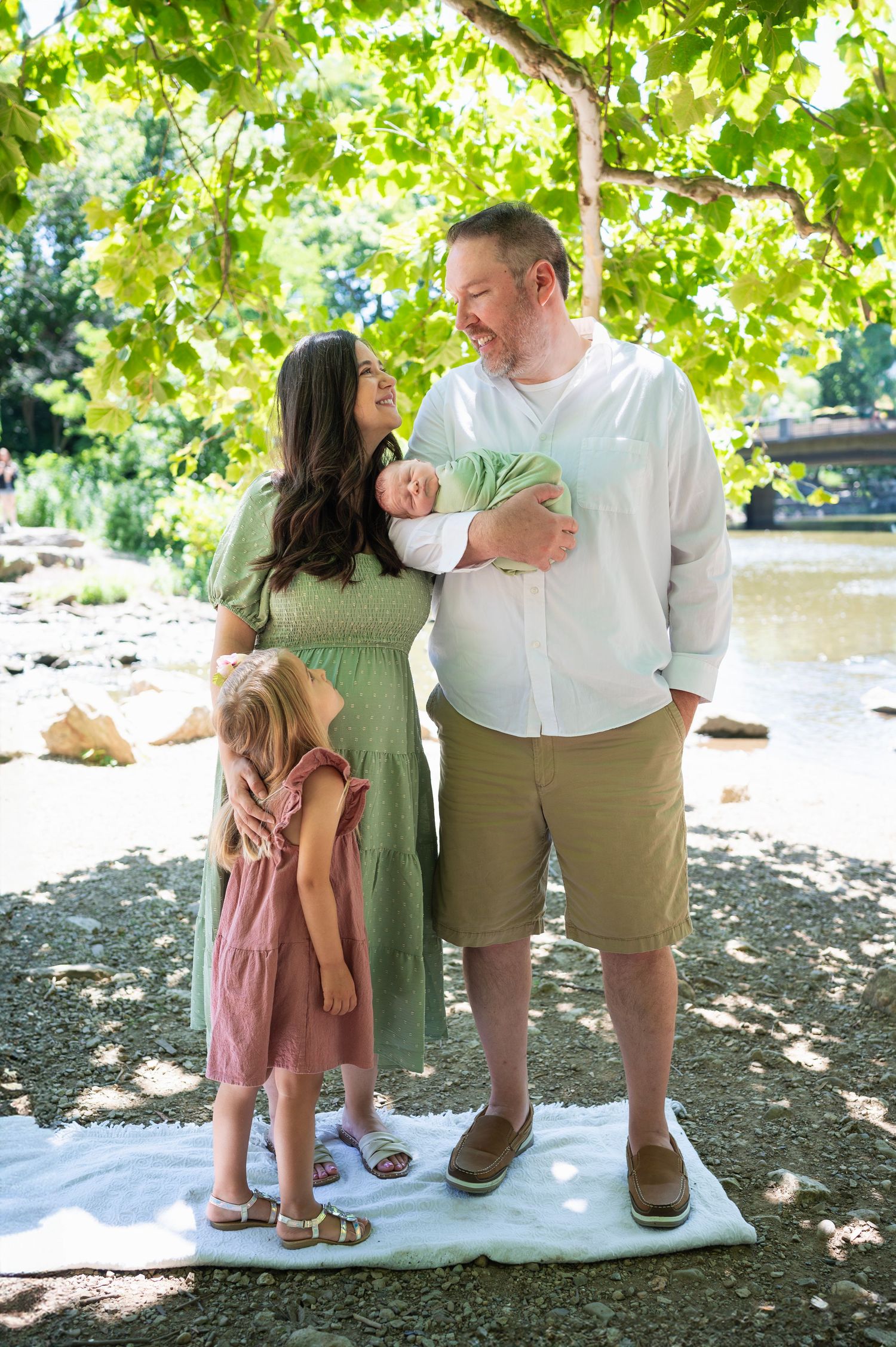 Family of four standing on a blanket by the river, parents smiling at each other while holding newborn, older daughter looking up at her dad