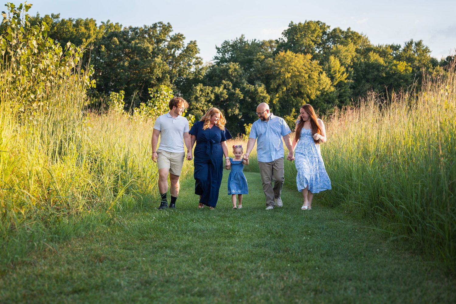 Family of five walking together through tall grass at golden hour, wearing blue and neutral tones.