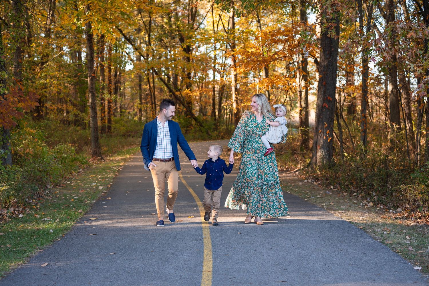 Family of four walking on a paved path lined with colorful autumn trees, parents holding kids’ hands. Rocky Fork Metro Park