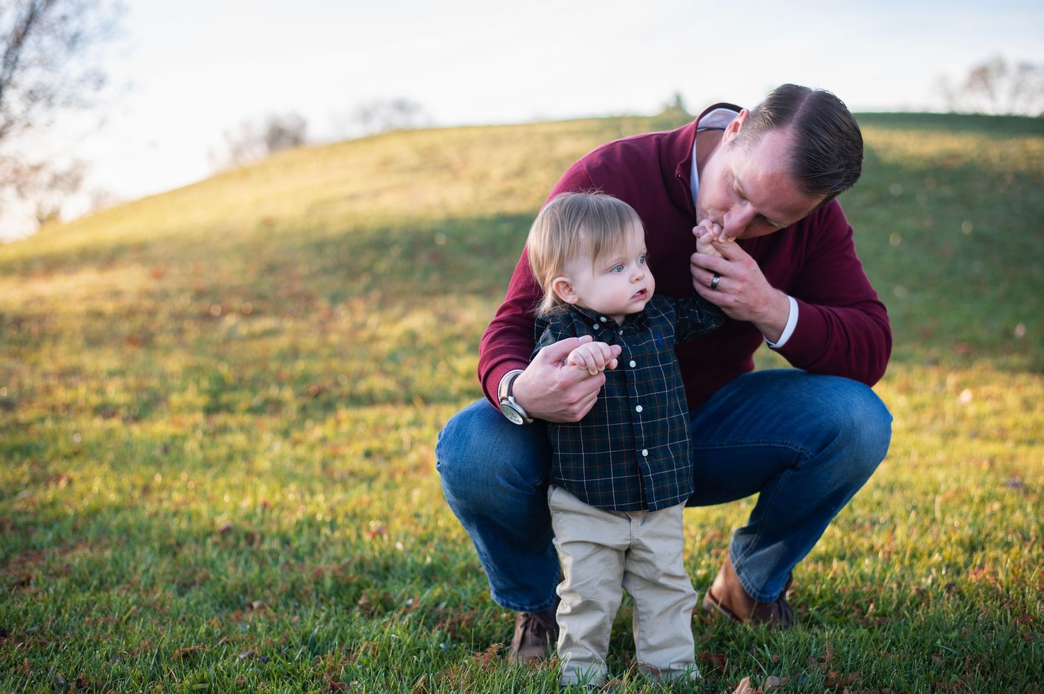 Father kneeling to kiss his toddler’s hand in a grassy park on a sunny autumn day. Highlands Aquatic Center