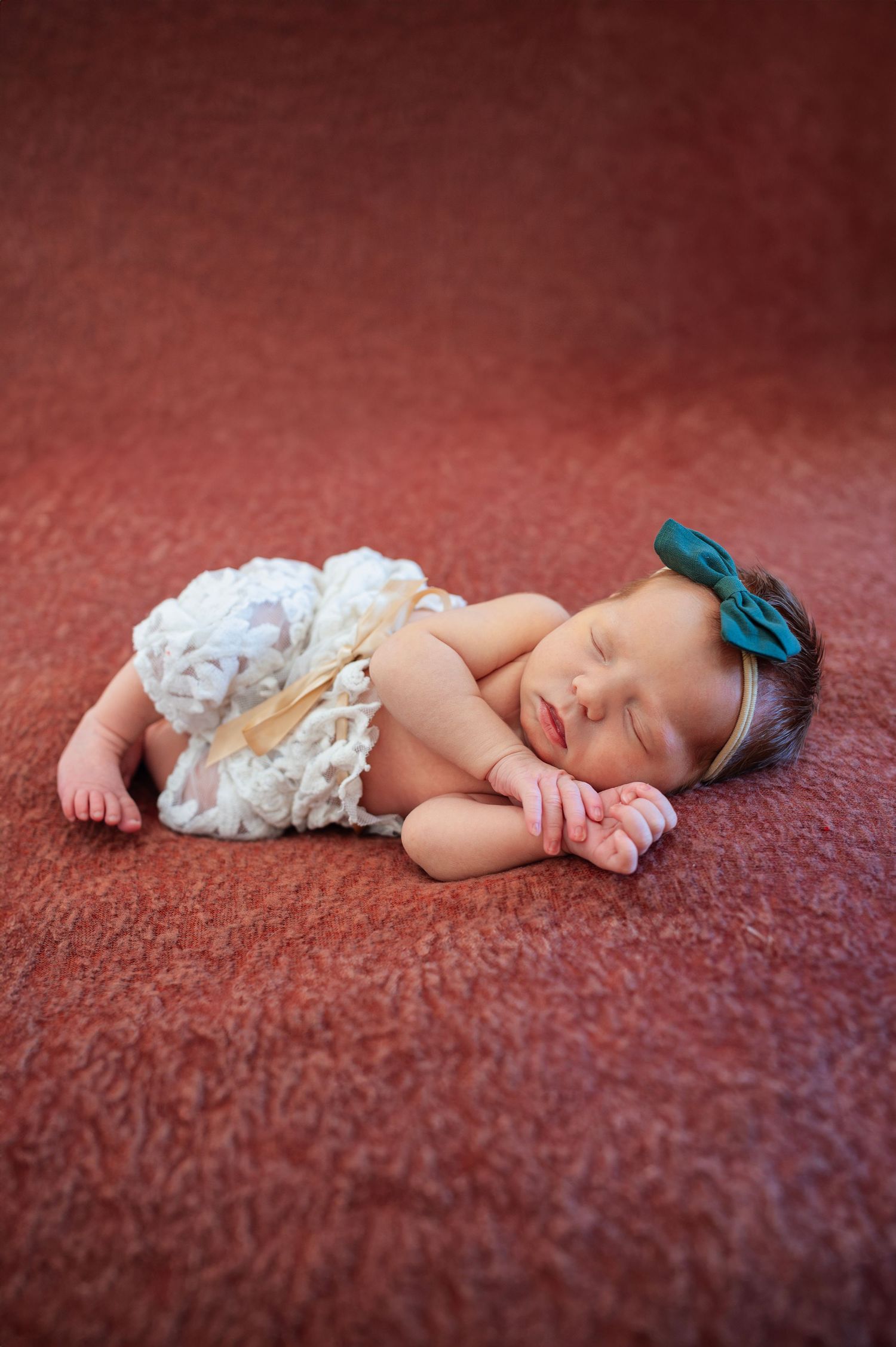 Newborn girl sleeps peacefully on rust-colored blanket, wearing lace pants and a dark green bow.